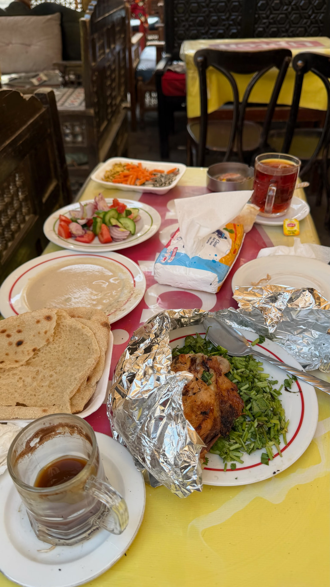 A café table laid with grilled chicken in foil, flatbread, tahini, salad and glasses of tea