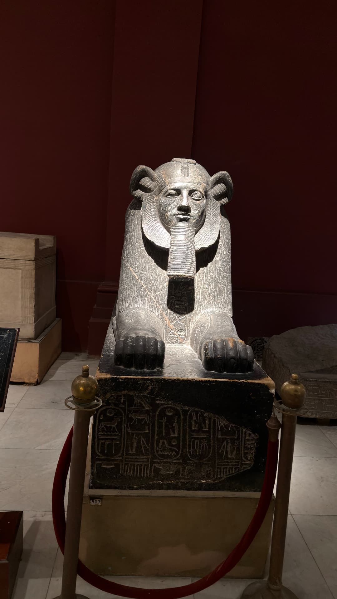 A dark stone sphinx with a pharaoh's face on an inscribed base behind a rope barrier