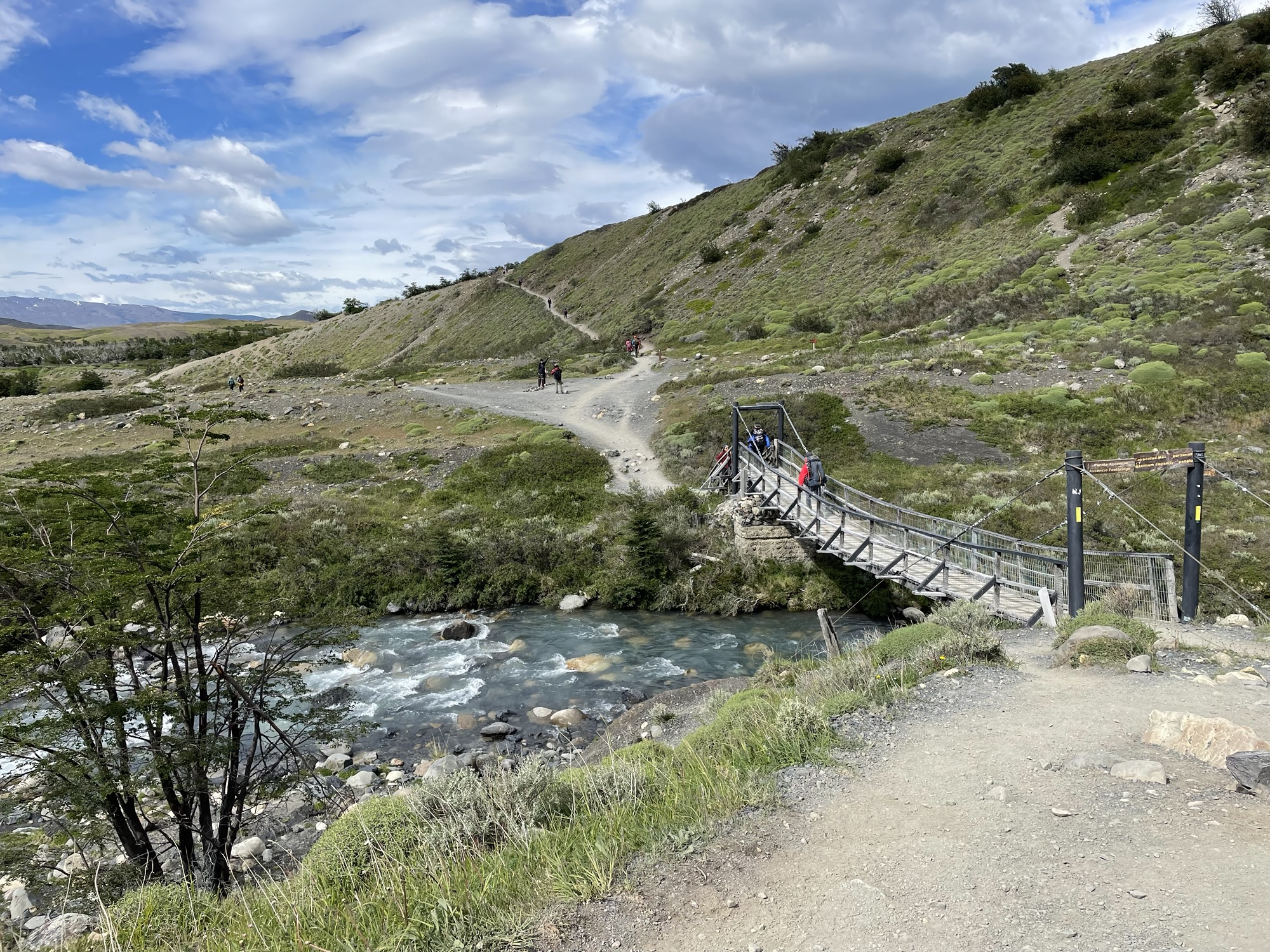 A small suspension footbridge crossing a churning river in a scrub-covered valley, with hikers crossing and distant hills