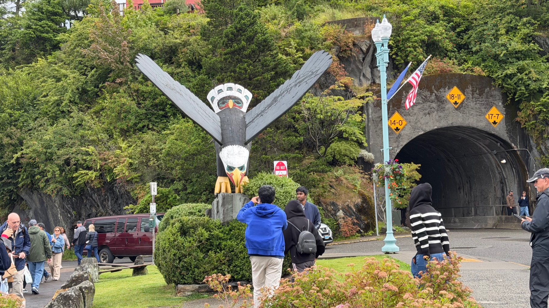 A carved eagle totem with outstretched wings standing beside a rock tunnel entrance in Ketchikan, with visitors taking photographs