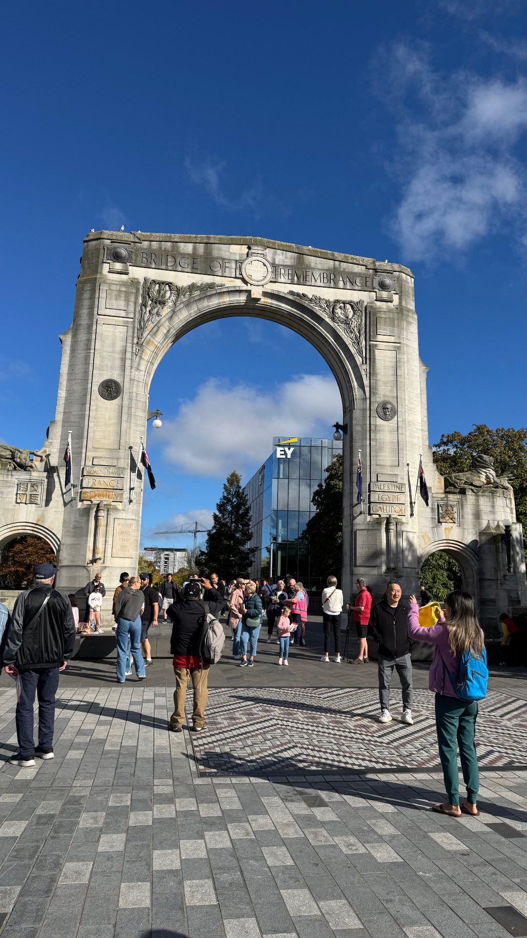 The stone Bridge of Remembrance arch in Christchurch under a blue sky, with visitors gathered on the paved square in front of it