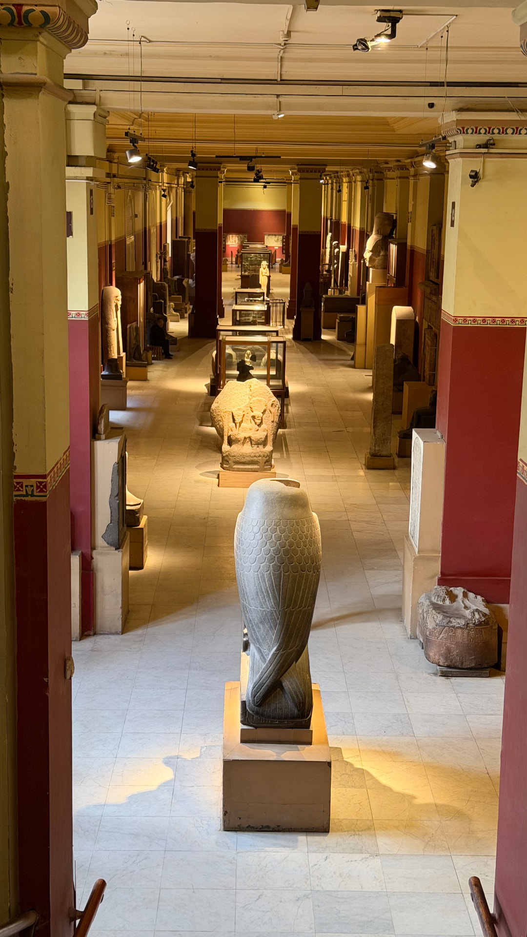 A long museum hall lined with columns and statues, a granite falcon on a plinth in the foreground