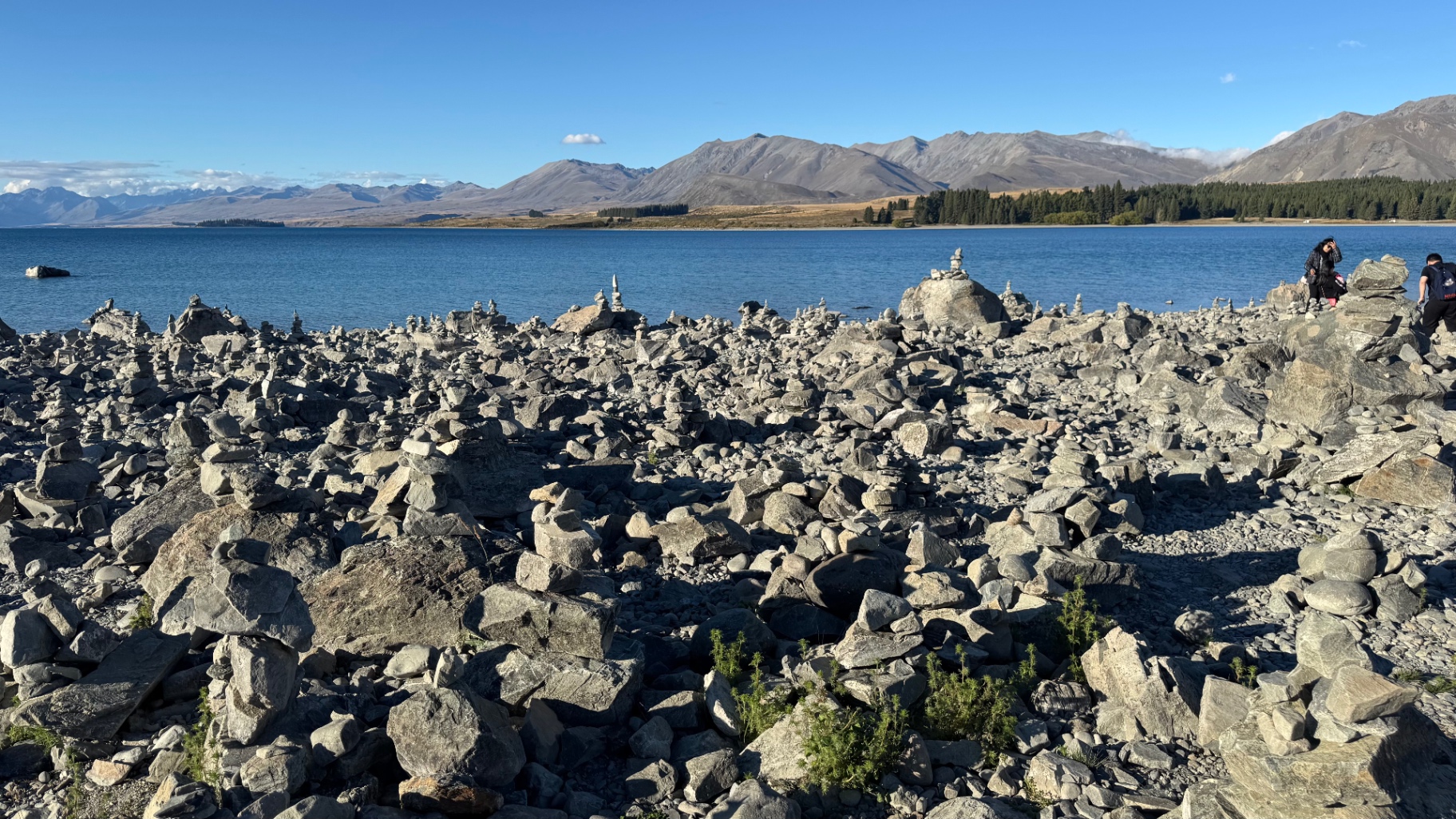 A rocky lake shoreline covered with hundreds of stacked stone cairns, with mountains beyond