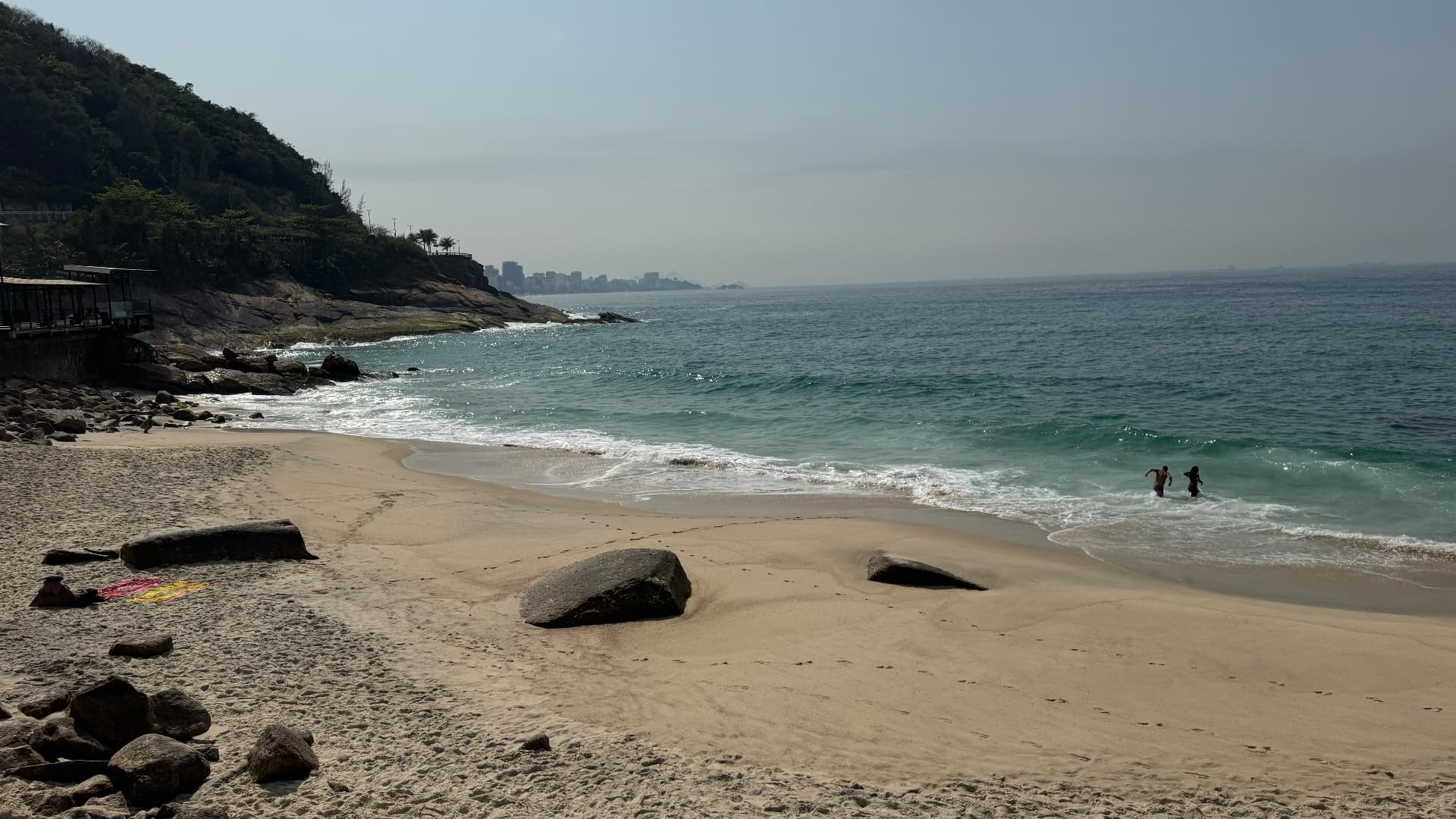 Quiet stretch of sand with large boulders, breaking waves, and two people wading
