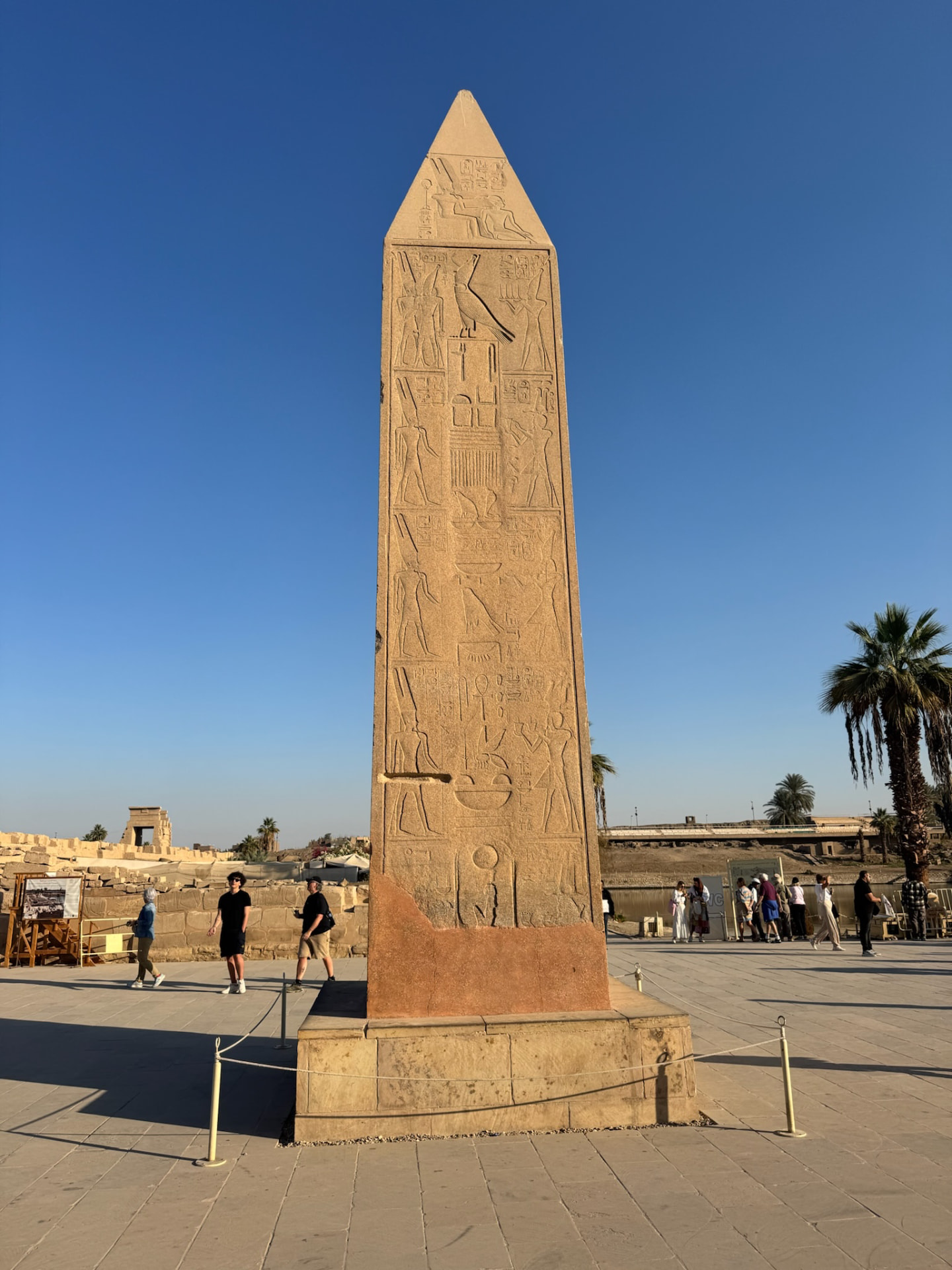 A carved obelisk standing on a paved court at Karnak with palms and visitors behind