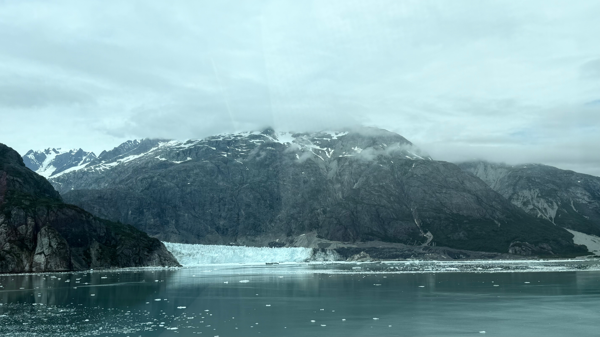 A tidewater glacier beneath a mountain wrapped in low cloud, with ice fragments across the bay