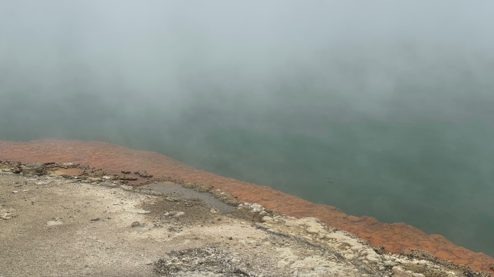 The orange mineral rim of a steaming green thermal pool half hidden by mist