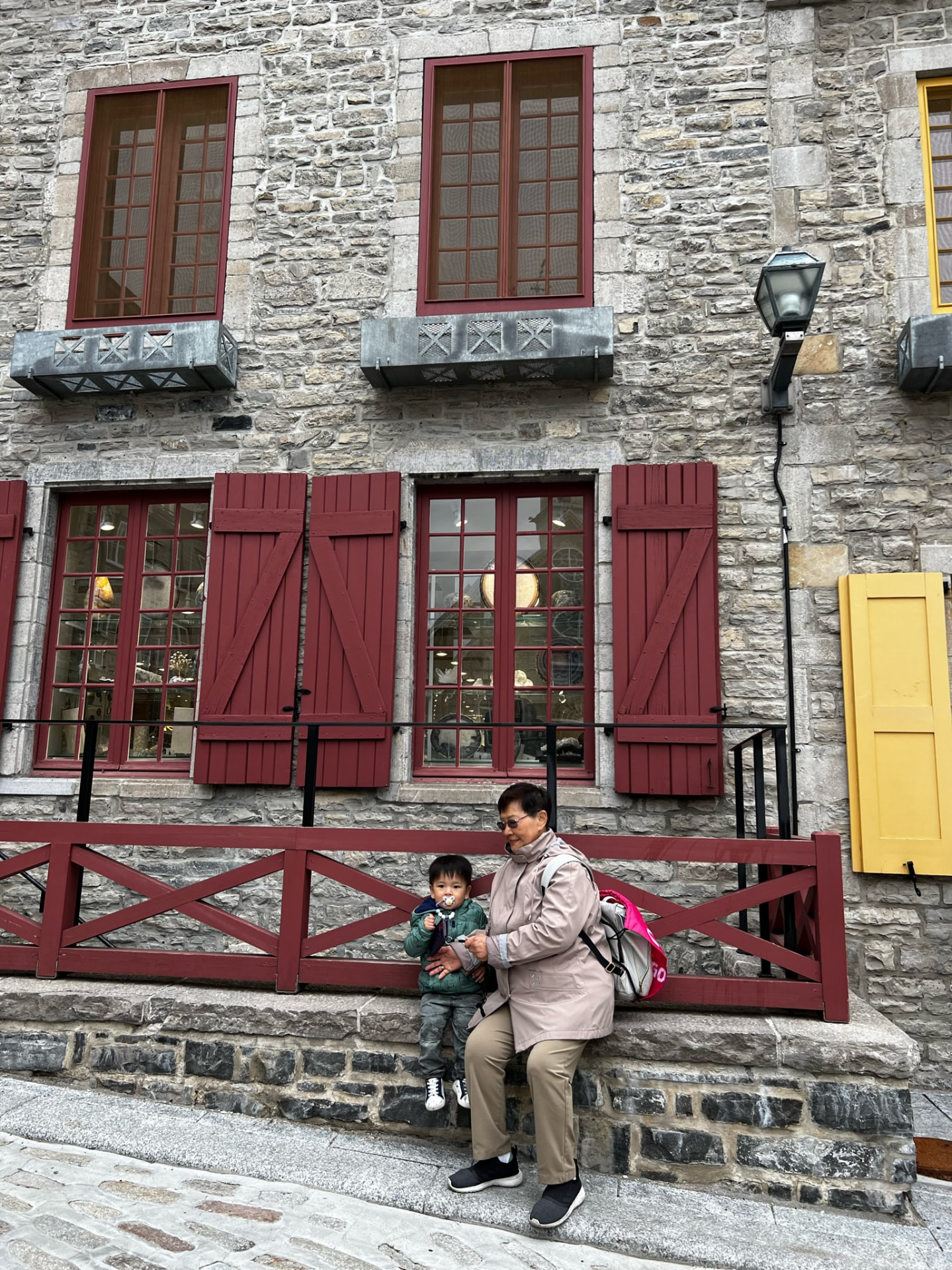Grandmother and child sitting by a stone building with red shutters