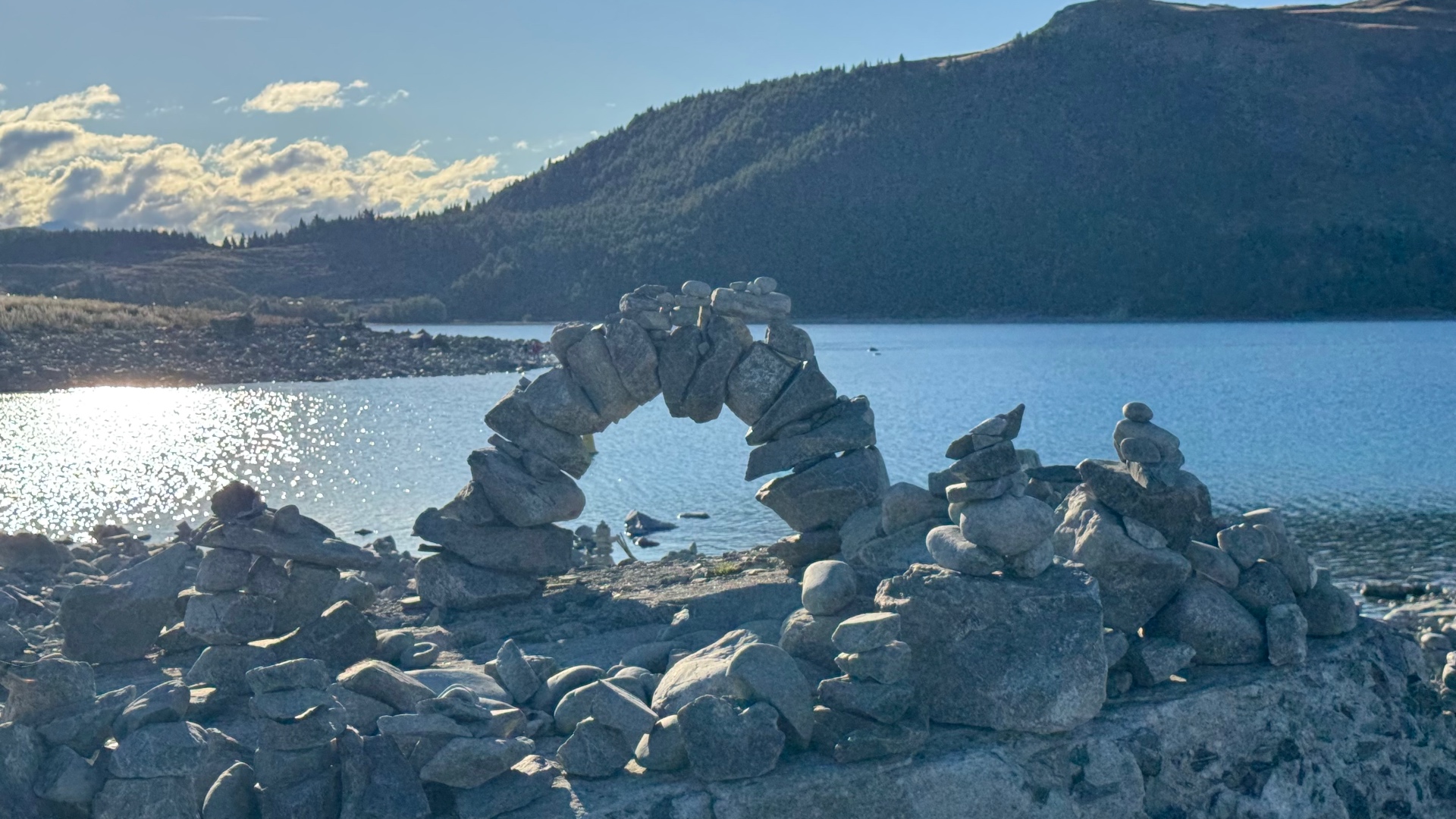 An arch built from stacked stones on the lake shore, backlit by sunlight on the water