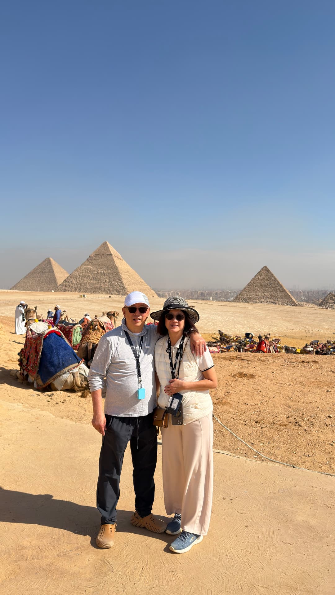 Ken and Becky standing together on the Giza plateau with three pyramids and resting camels behind them