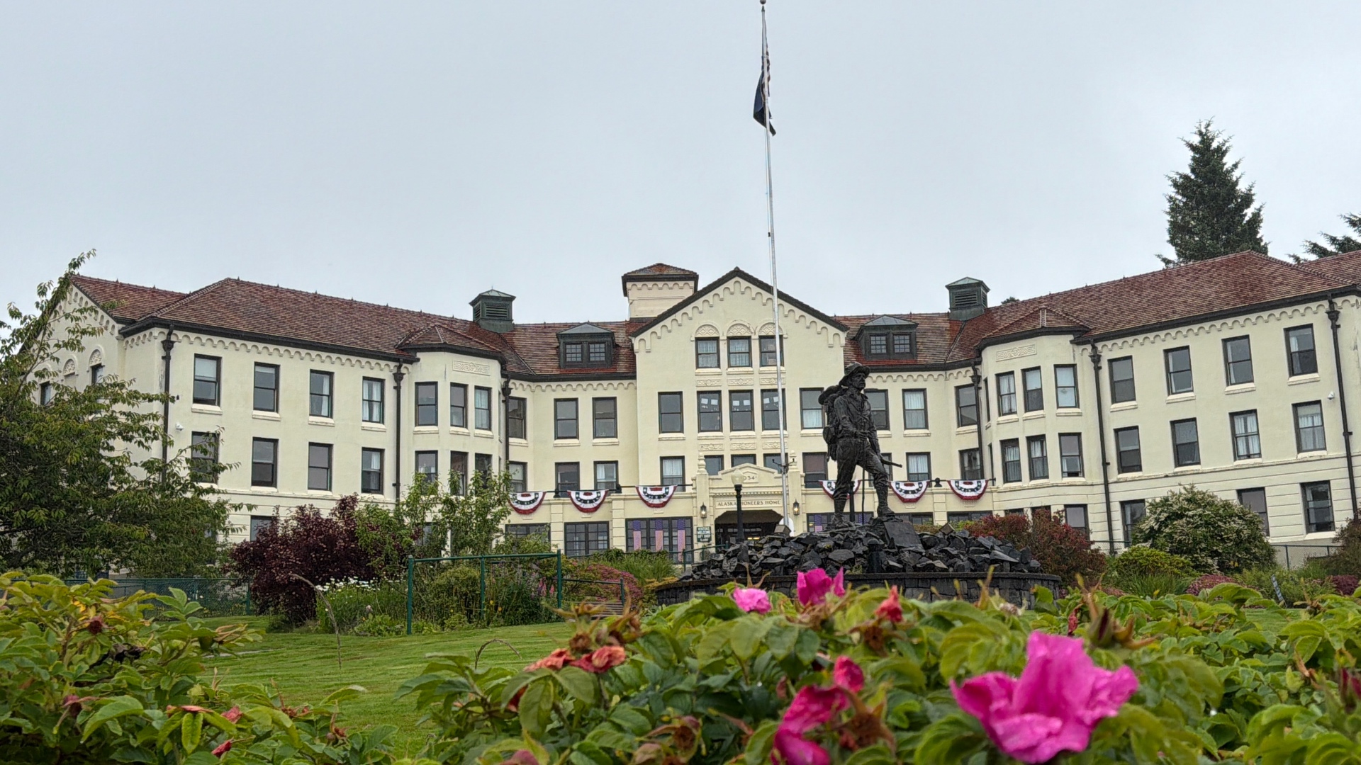 The Alaska Pioneers Home, a large cream building with a red tile roof, seen across a lawn with a bronze prospector statue and pink roses in the foreground