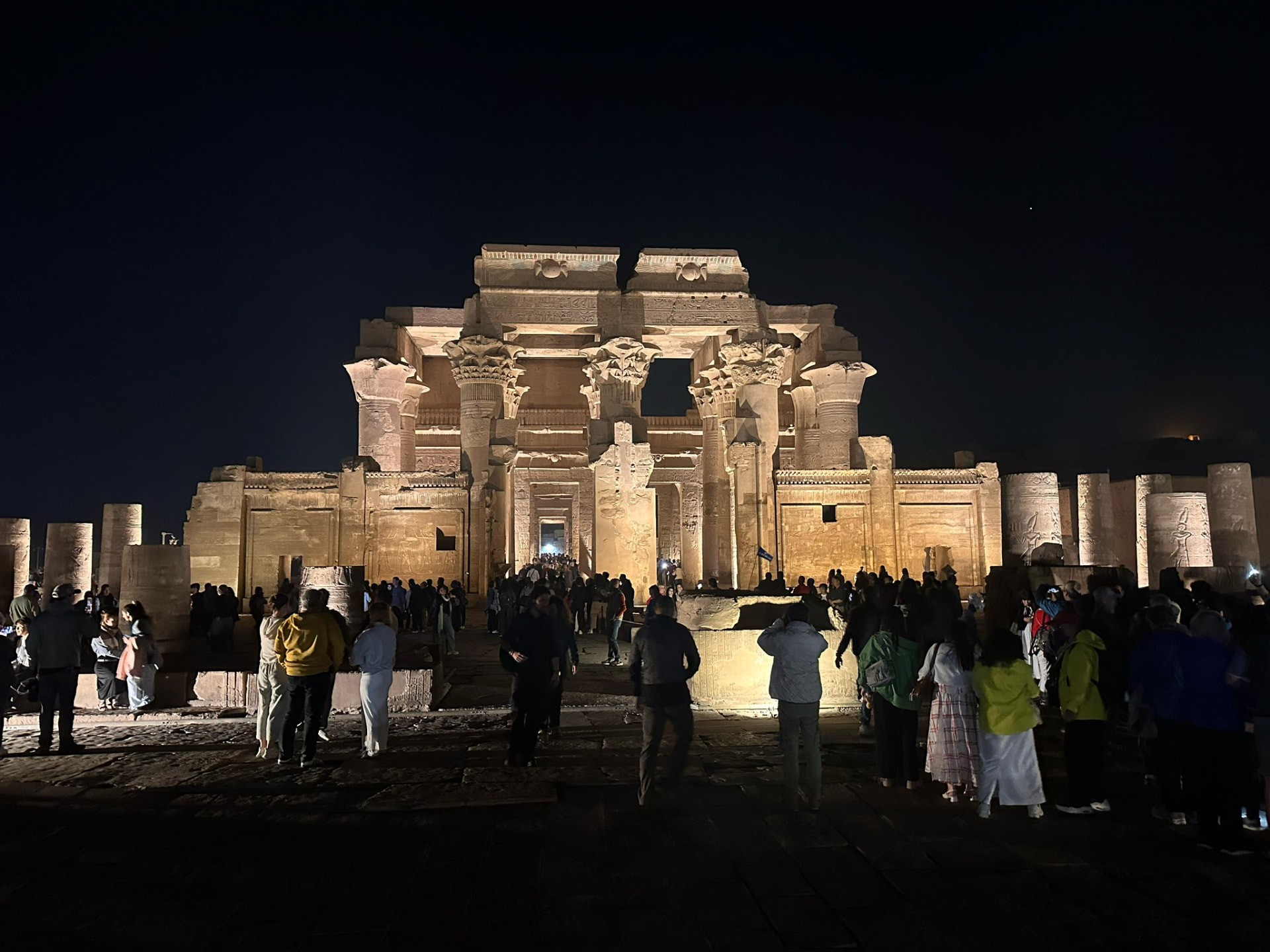 The full facade of the Temple of Kom Ombo floodlit at night with a crowd gathered in front