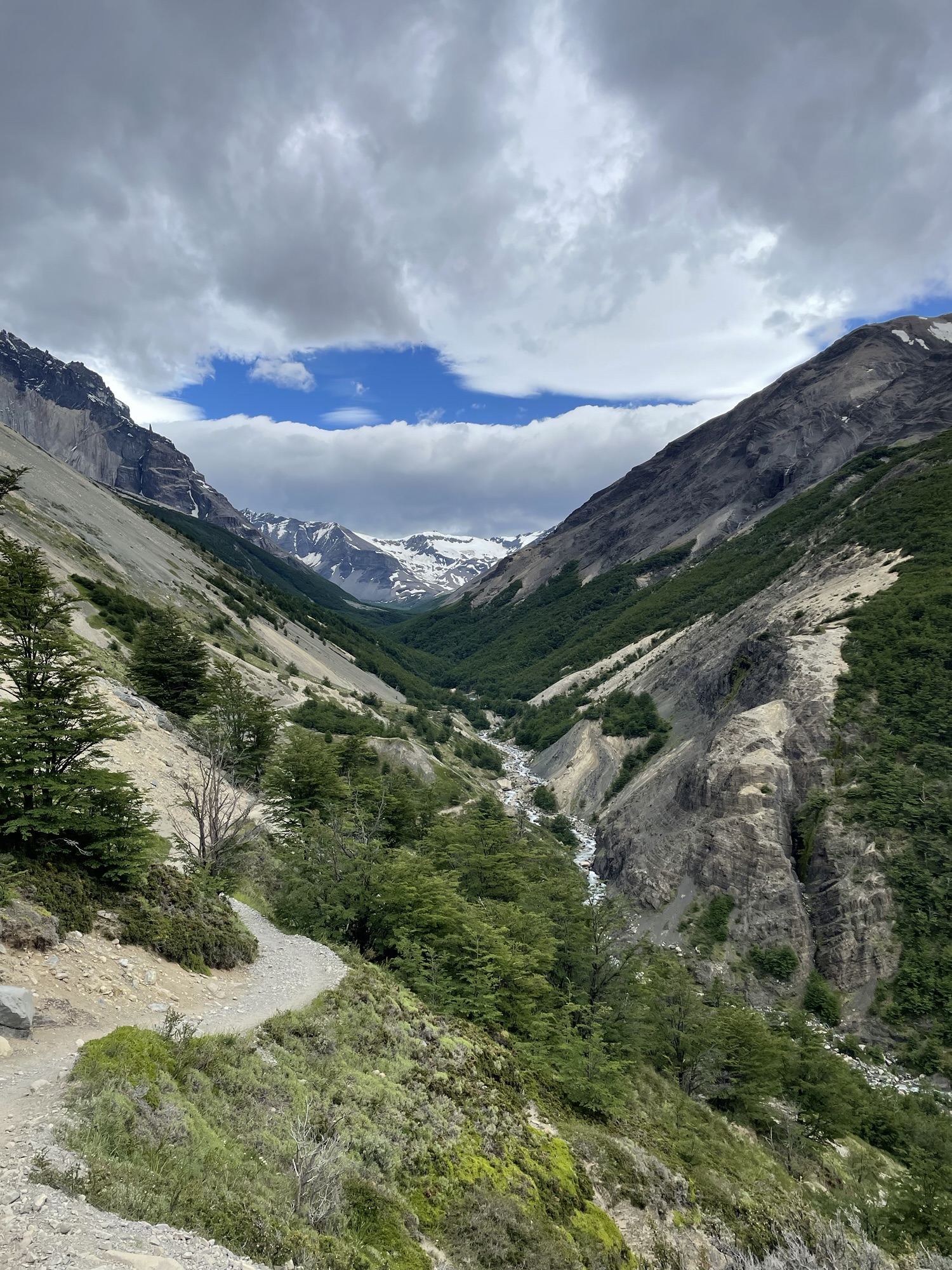 A vast V-shaped mountain valley seen from a high hiking trail, with a thin river winding along the floor and snow-capped peaks under heavy clouds