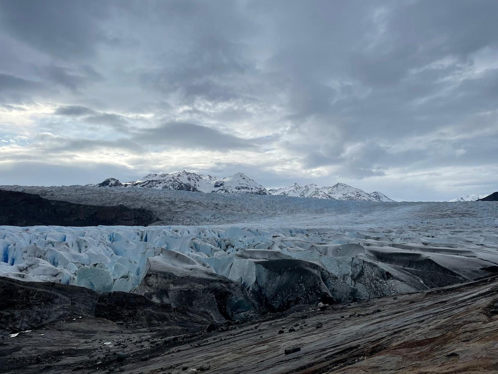A panoramic view of a glacier terminus rising as a jagged wall of luminous blue ice above dark striated rock, with snow-covered mountains beyond