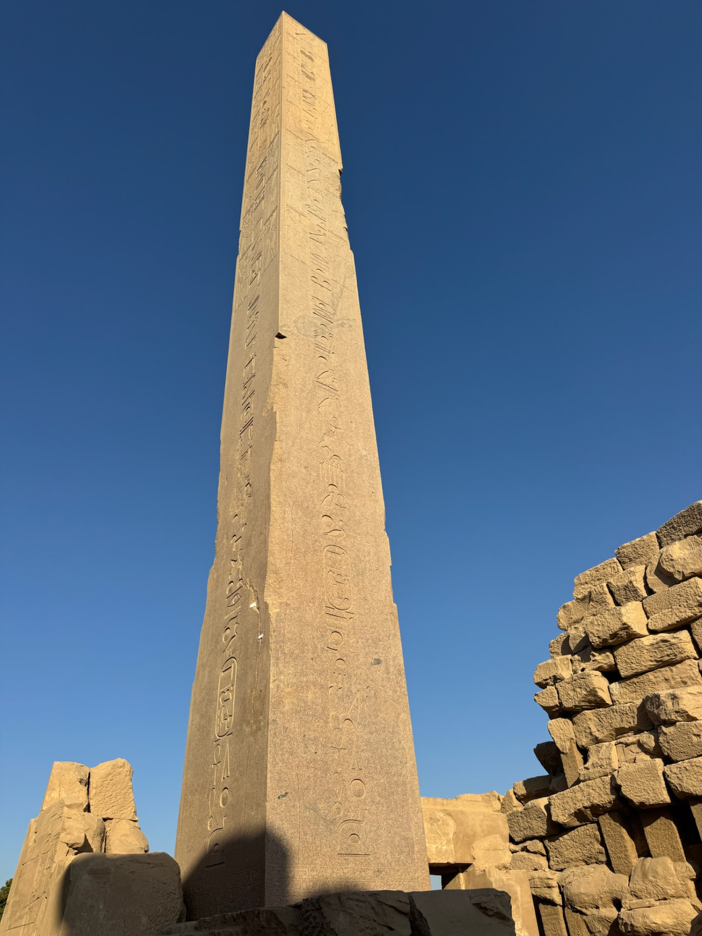 A granite obelisk at Karnak rising against a deep blue sky beside a ruined wall