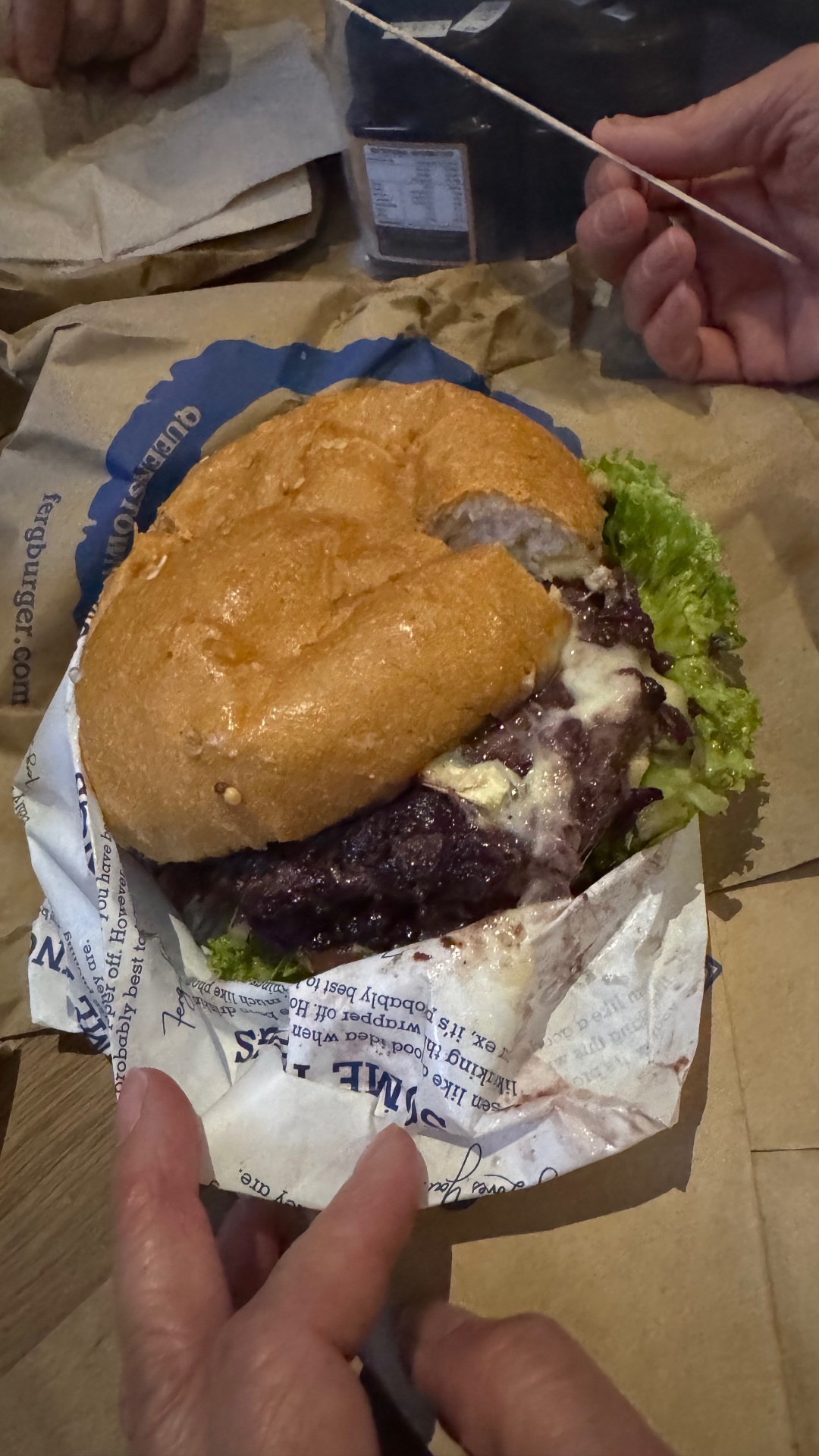 A close-up of a burger with melted cheese, lettuce, and dark sauce on Fergburger wrapping