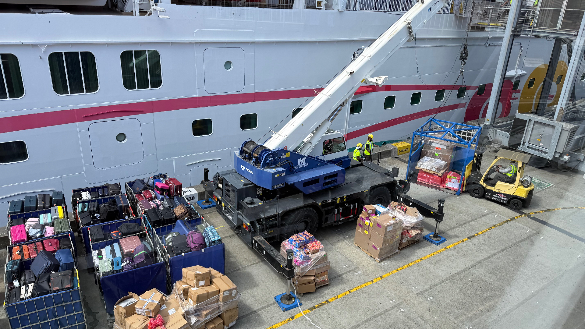 A blue and white crane loading luggage and cargo pallets onto a white cruise ship at the Seattle dock