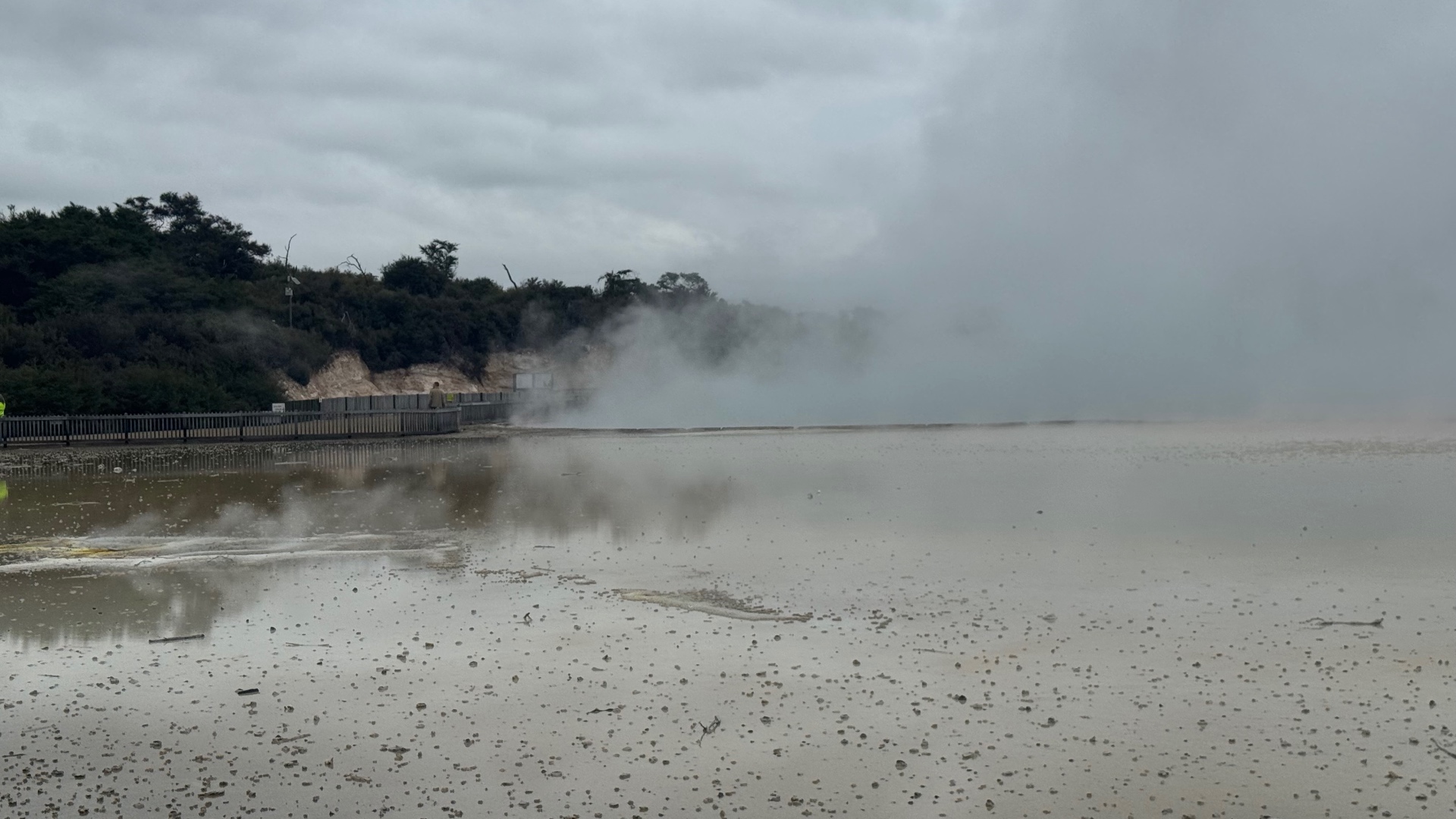 Steam drifting across a pale geothermal flat beside a fenced walkway under grey cloud
