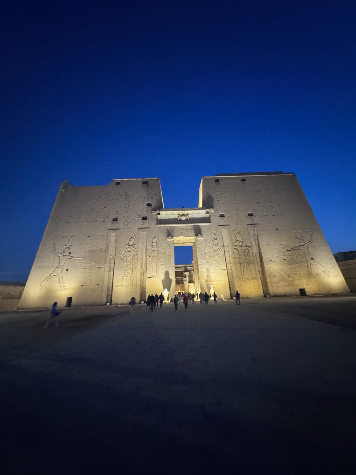 The floodlit pylon of the Temple of Edfu at night with visitors walking toward the lit doorway
