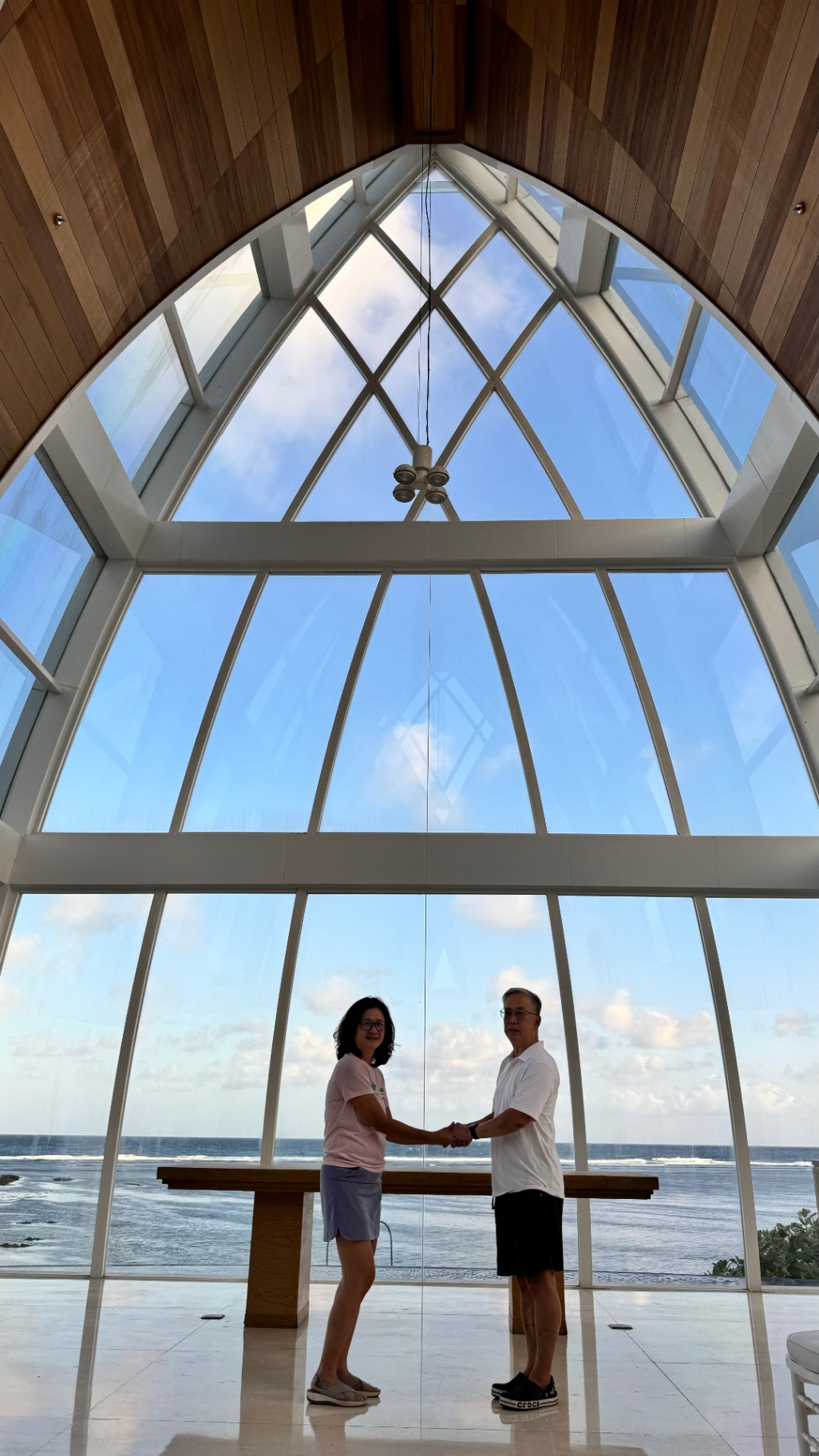 Becky and Ken holding hands beneath the chapel's soaring window overlooking the sea