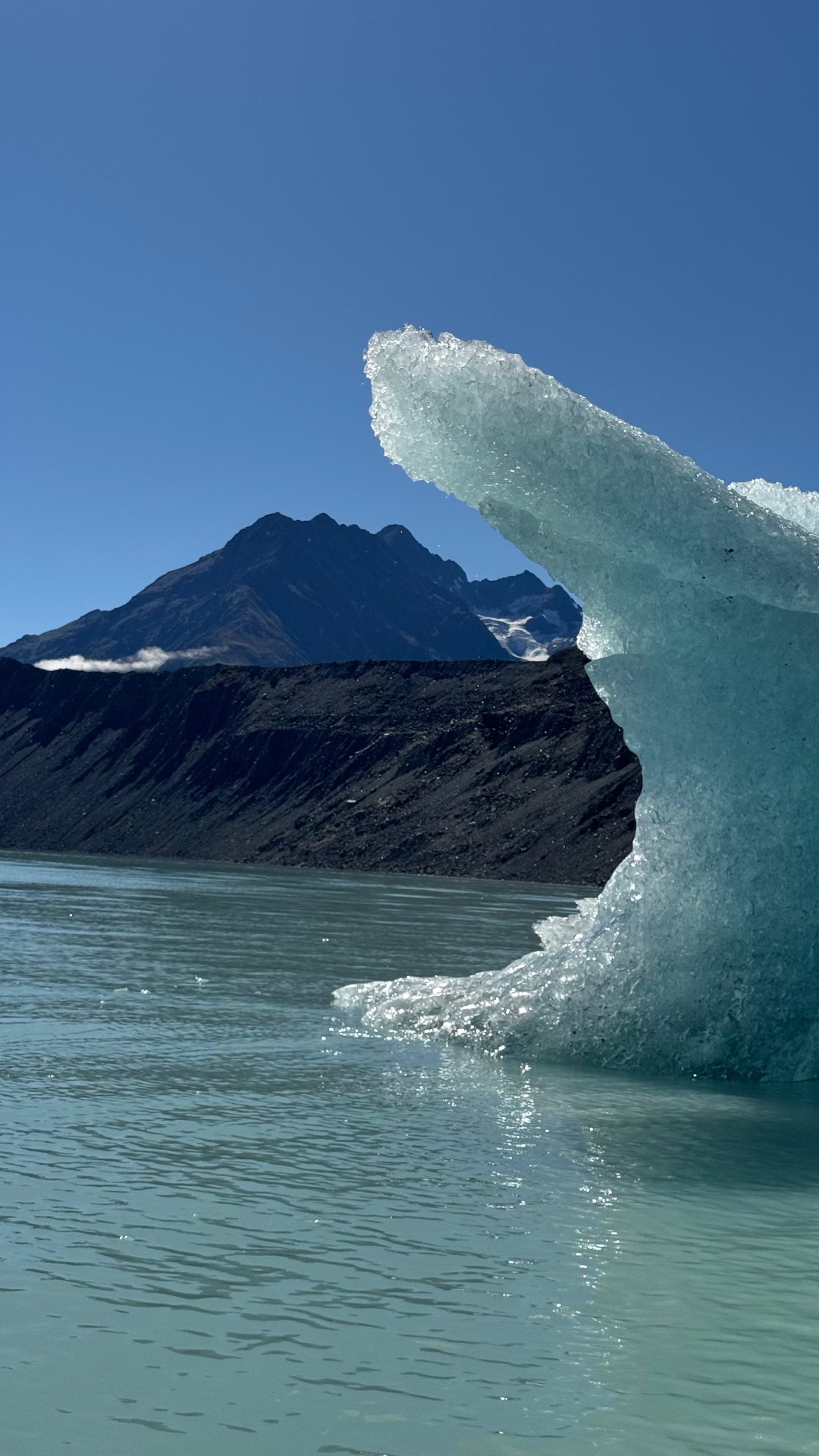 A close view of a crystalline blue-green iceberg edge rising out of the lake in front of a dark moraine wall