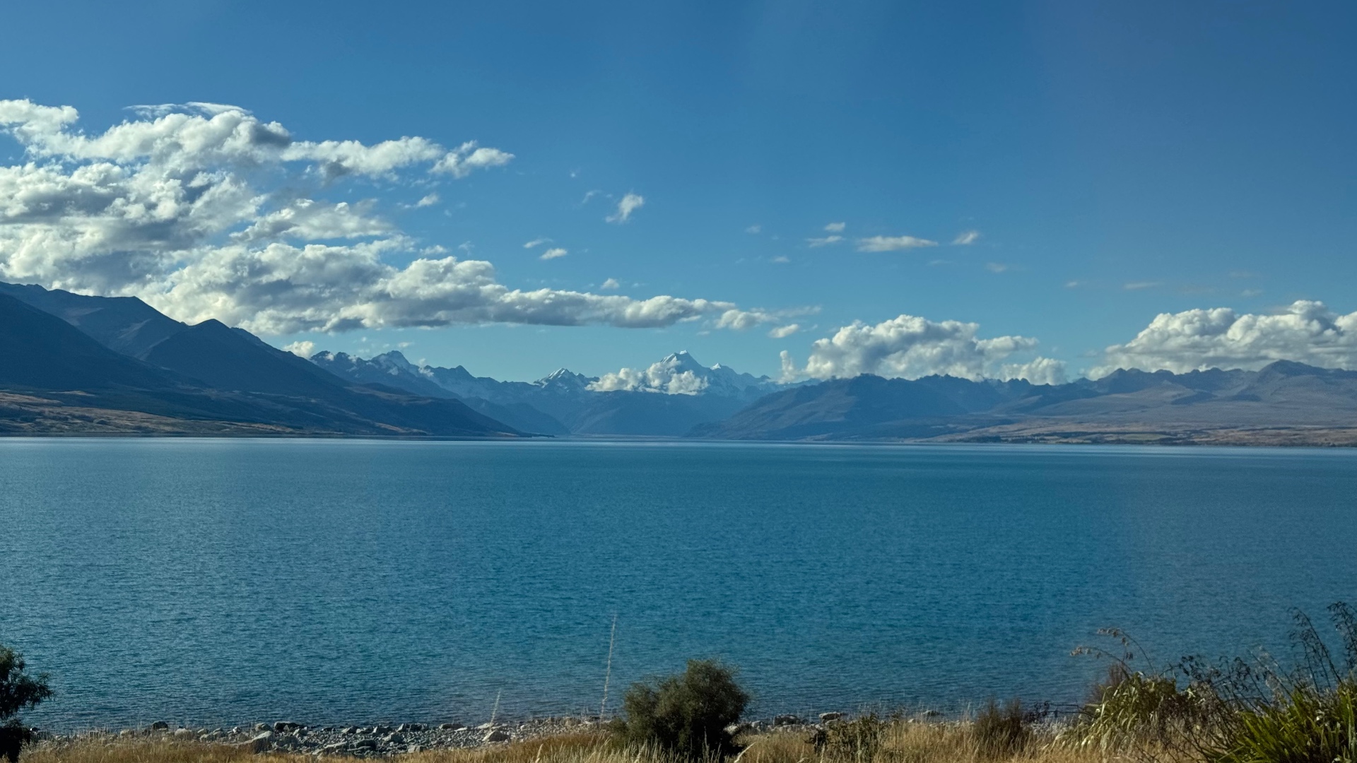 A wide blue alpine lake with snow-capped mountains, including Aoraki / Mount Cook, on the horizon under a bright sky