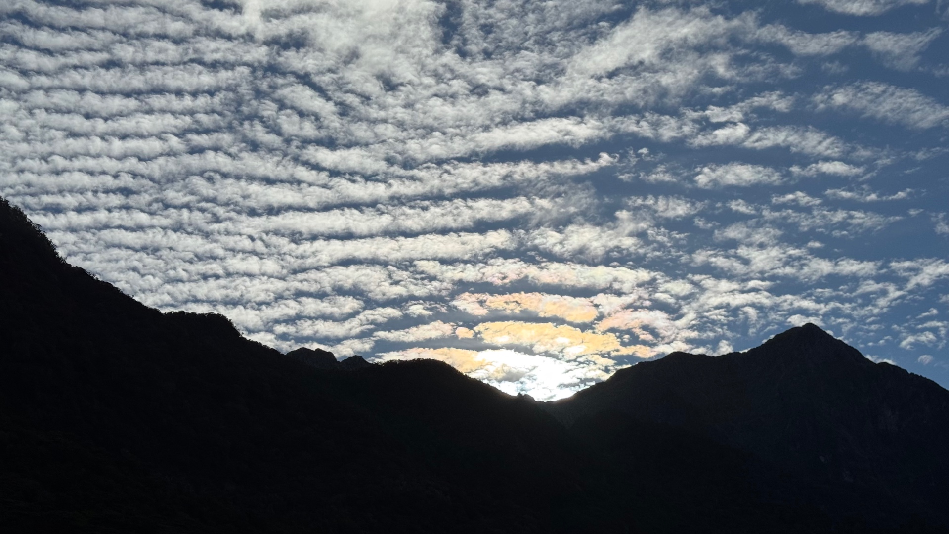 A striking sky of wavy banded clouds with iridescent light above the black silhouette of mountain ridges