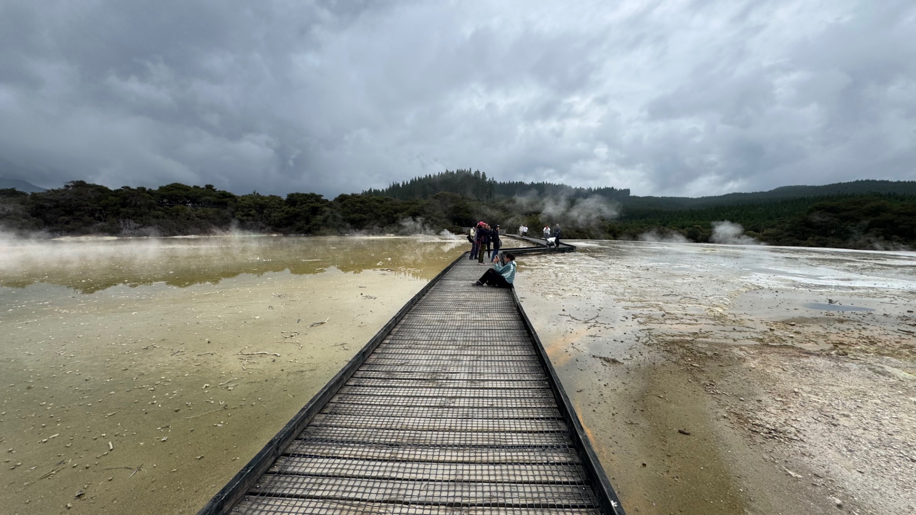 A long boardwalk running straight into a wide steaming geothermal basin under heavy clouds