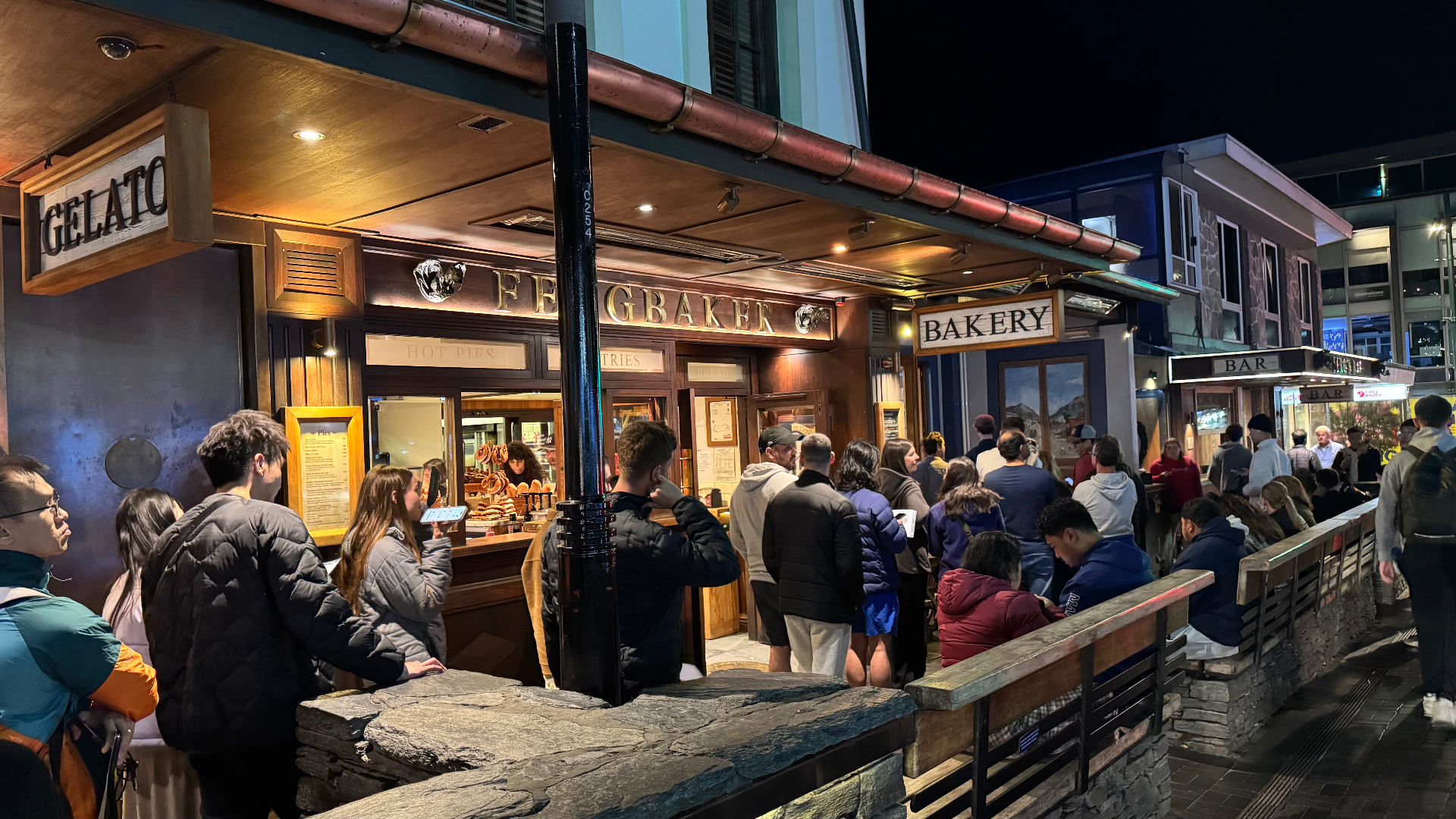 A nighttime street view of the Fergbaker storefront in Queenstown, with a long line of people waiting outside