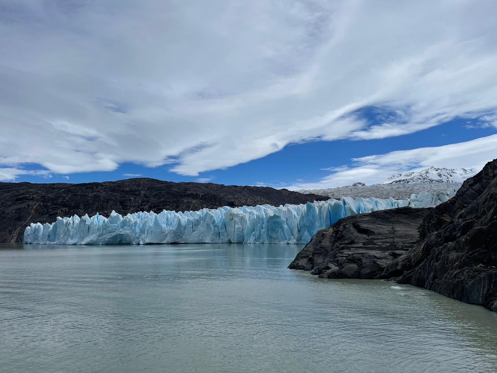 A glacier meeting a milky green-grey lake, its jagged blue and white ice wall reflected in the still water under a partly cloudy sky
