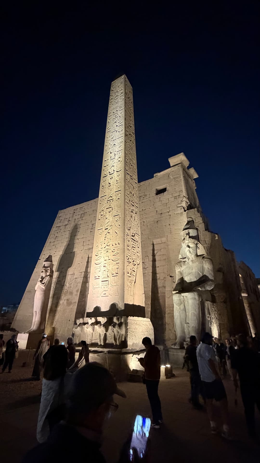 The Luxor obelisk lit gold at night beside a colossal seated statue of Ramesses