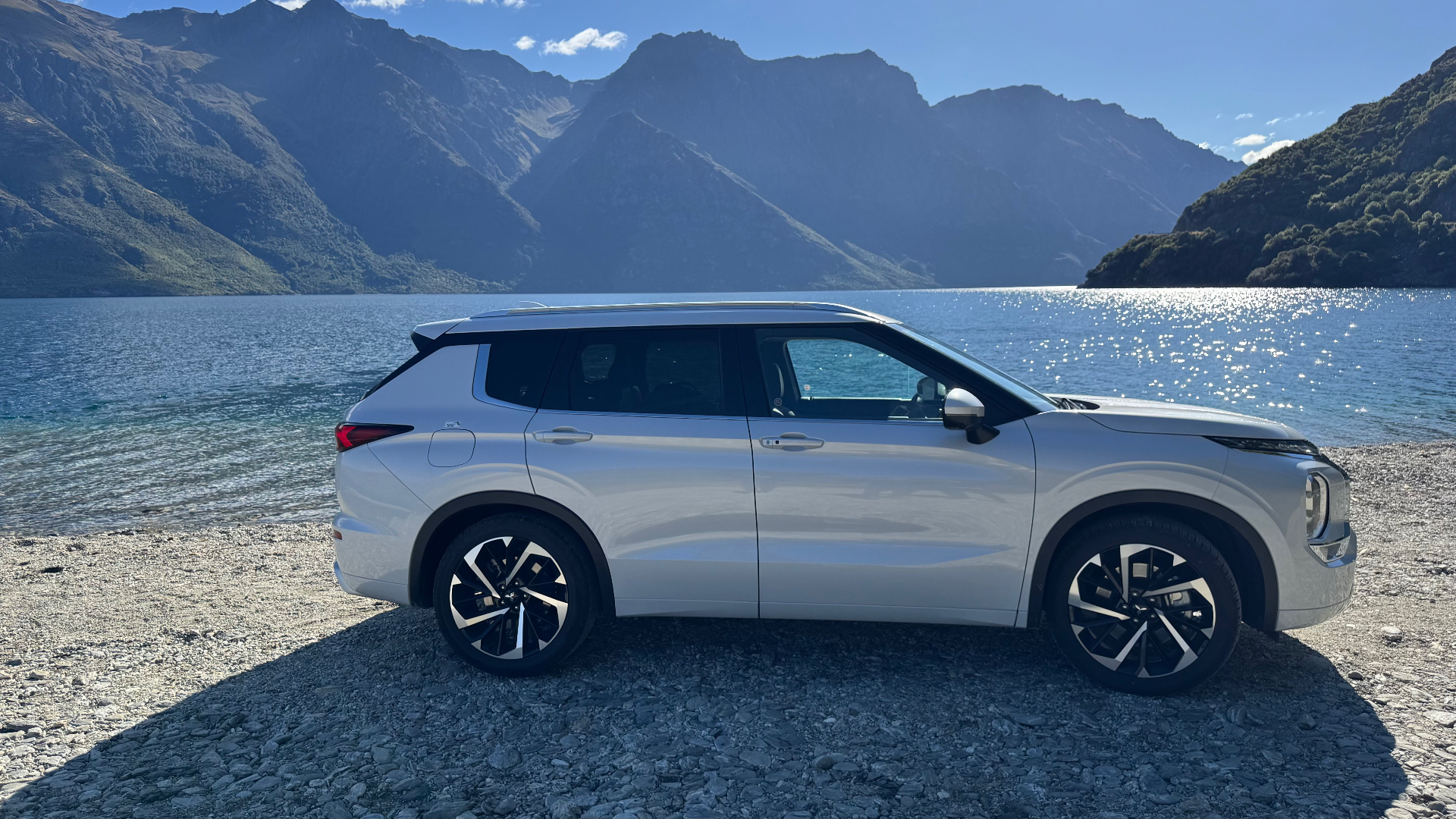 A white rental SUV parked on a stony lakeshore with mountains behind