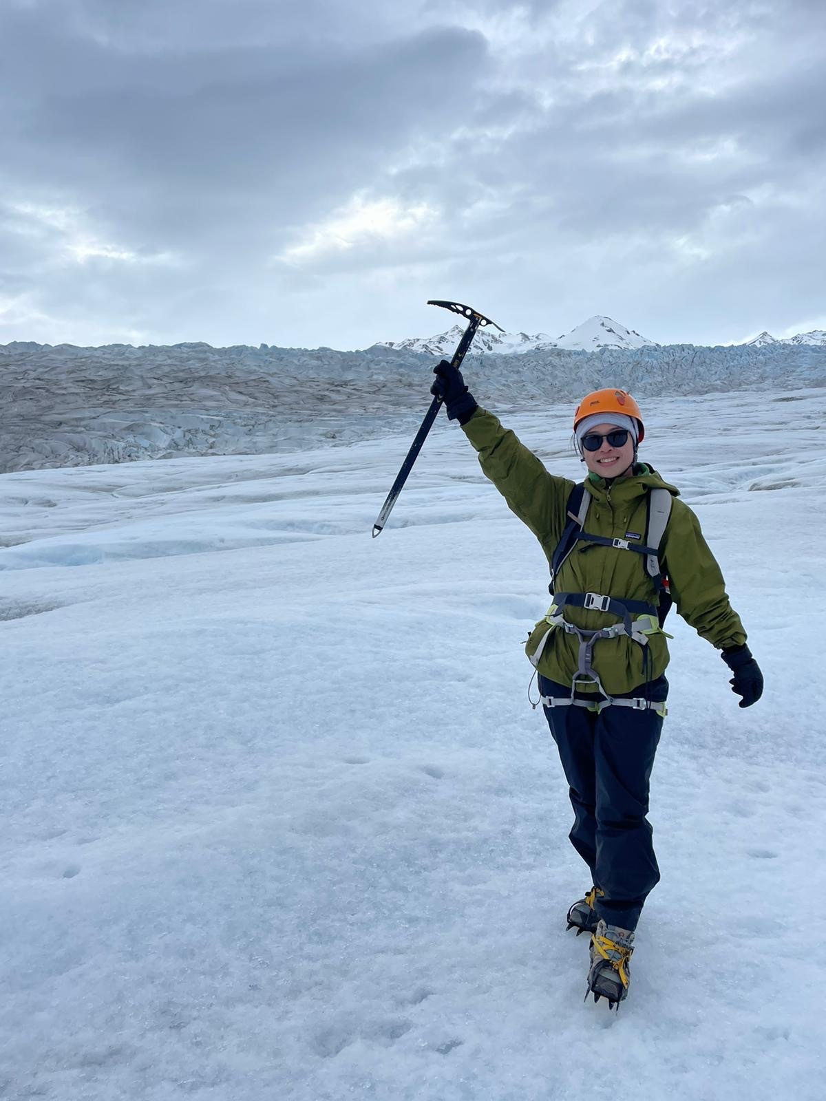 A person in mountaineering gear stands triumphantly on a glacier holding an ice axe aloft, smiling beneath an overcast sky