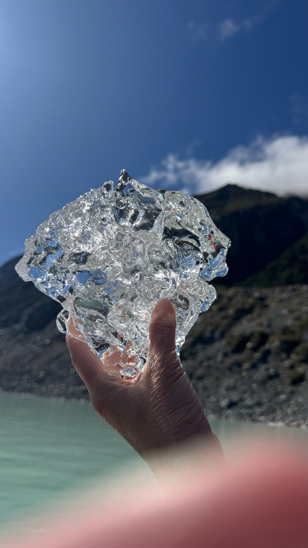 A hand holding up a clear chunk of glacier ice against the sun and blue sky