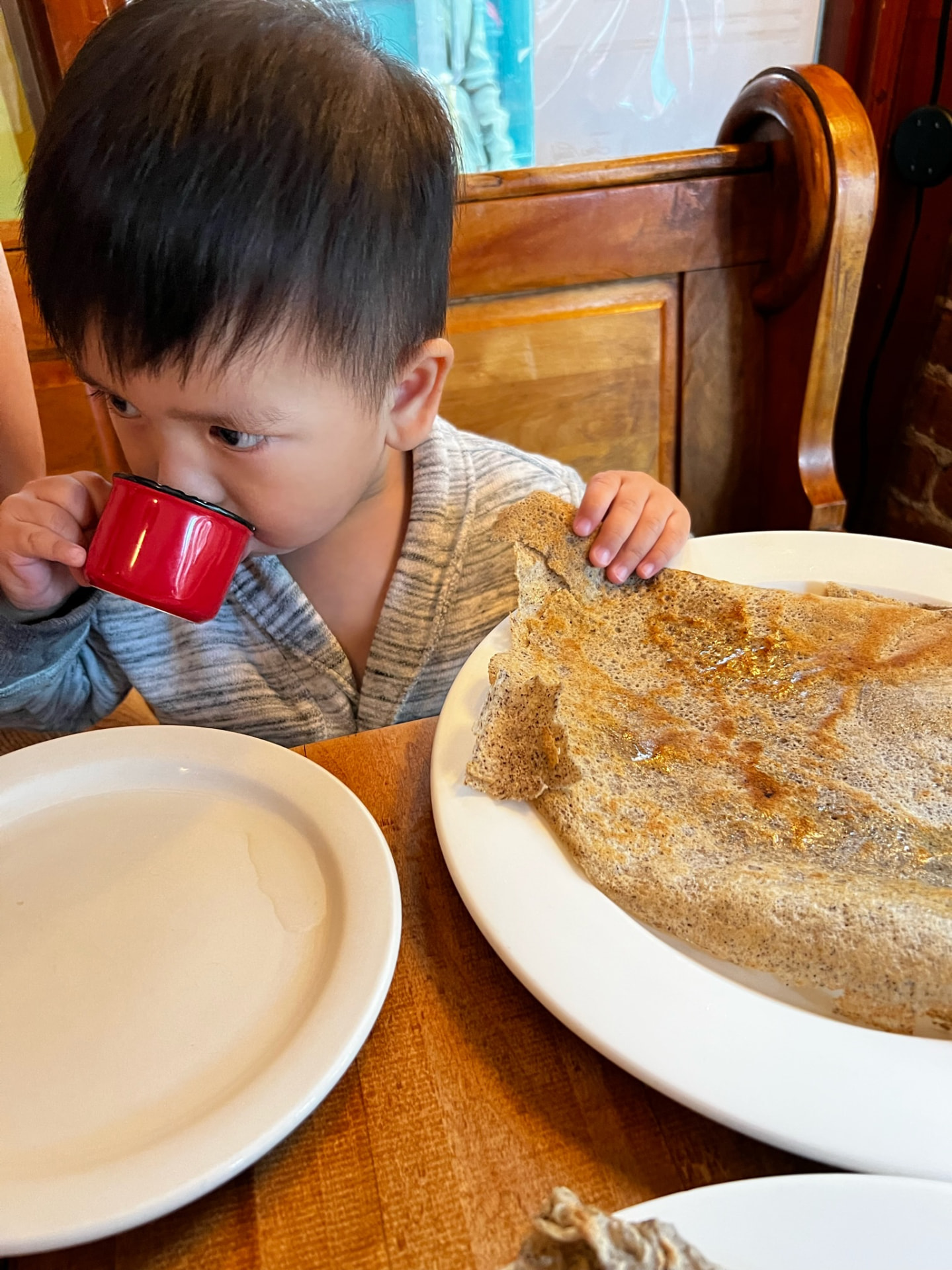 Young child drinking from a red cup beside a large buckwheat crêpe
