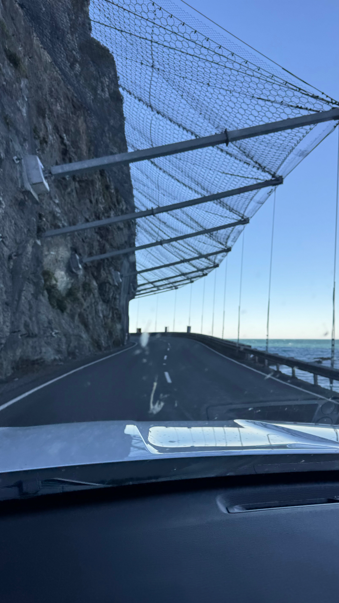 The coast road north of Kaikōura seen through a car windscreen, running under a rockfall net beside a cliff and the sea