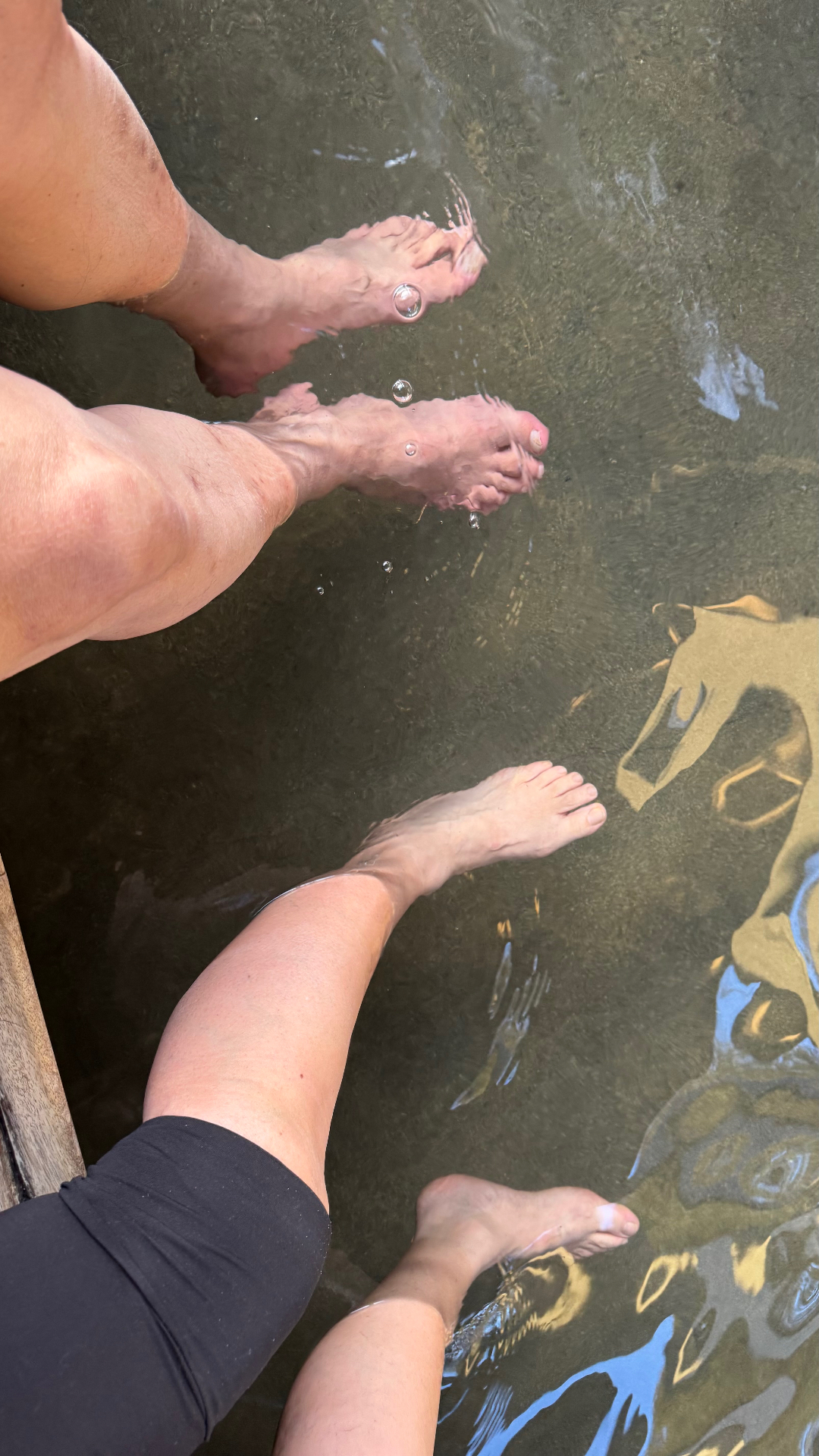 Four bare feet dangling into the warm, clear water of a natural hot stream from a wooden edge