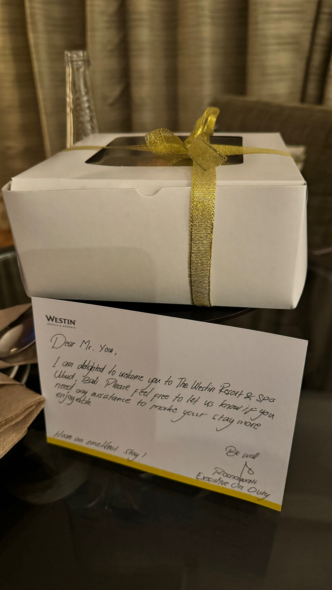 A boxed cake tied with gold ribbon beside a handwritten Westin welcome card addressed to Mr Yow