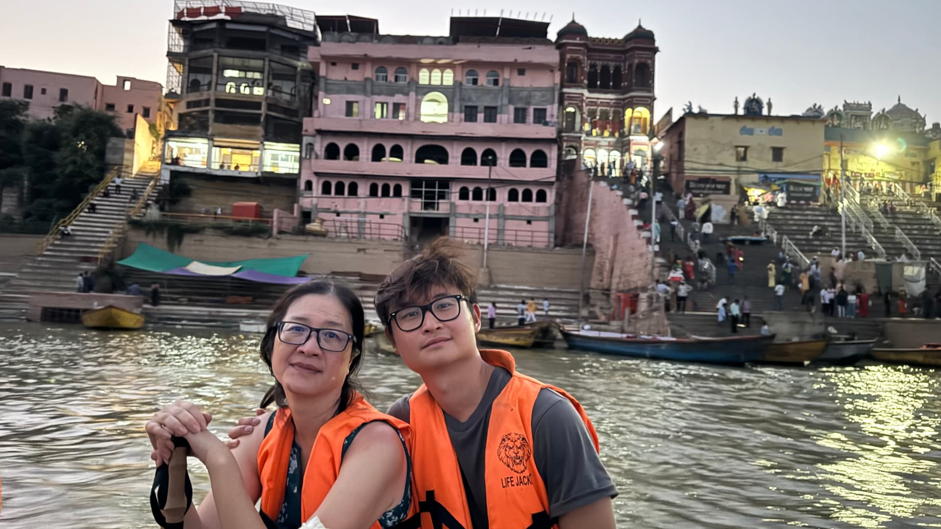 Two companions in life jackets pose with the waterfront behind them. — Varanasi, Uttar Pradesh, India.