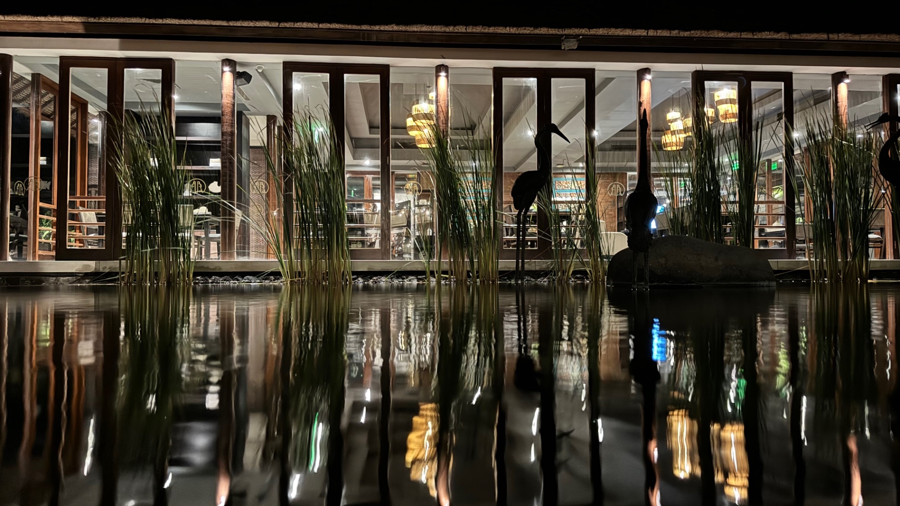 Heron sculptures and reeds silhouetted against the lit restaurant across the water