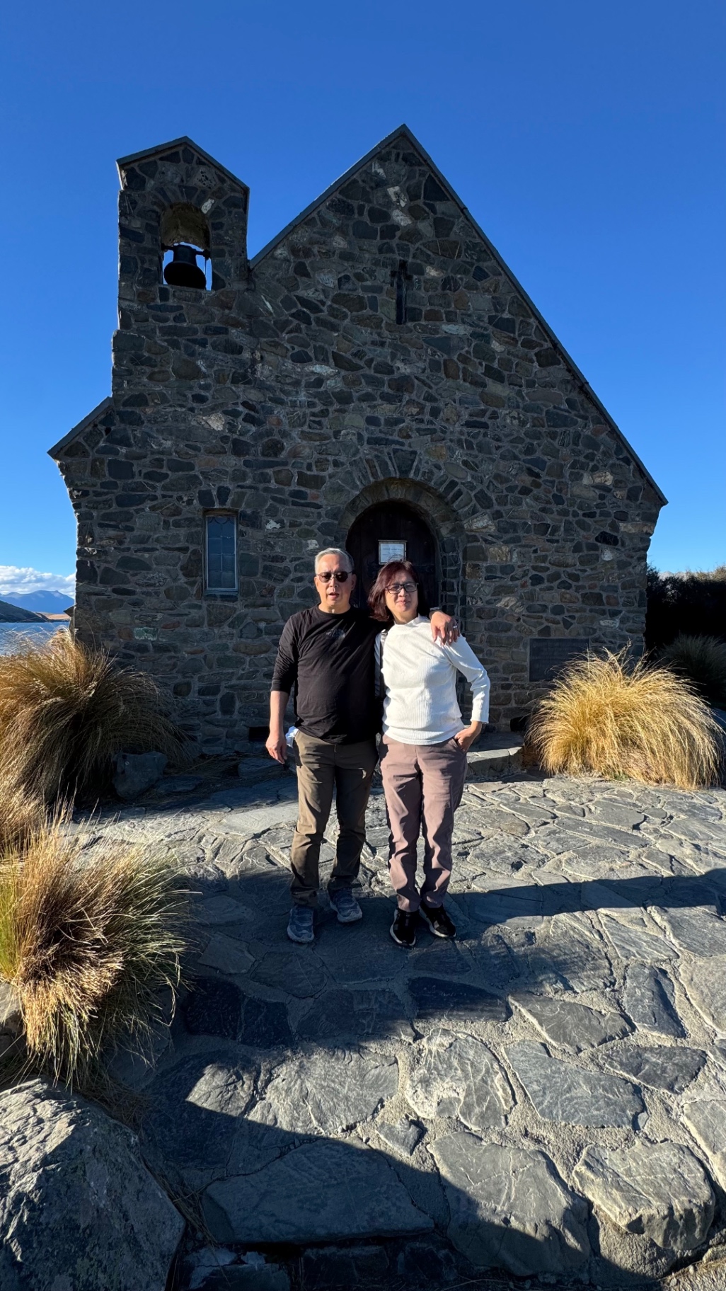 Two travellers standing together in front of the stone Church of the Good Shepherd under a deep blue sky