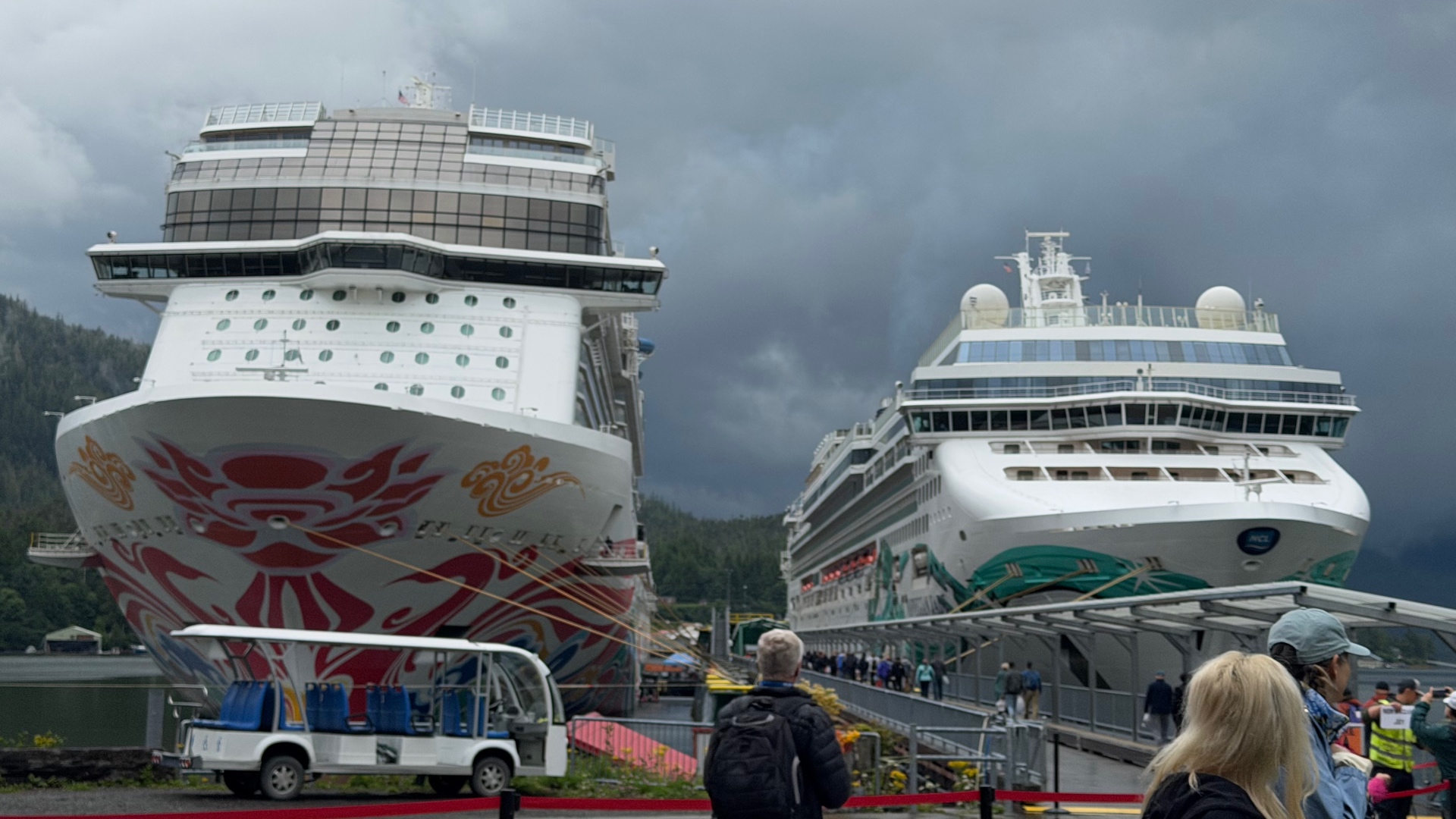 Two cruise ships berthed side by side at Ketchikan under heavy grey clouds, one with a large red and orange Native-inspired hull design