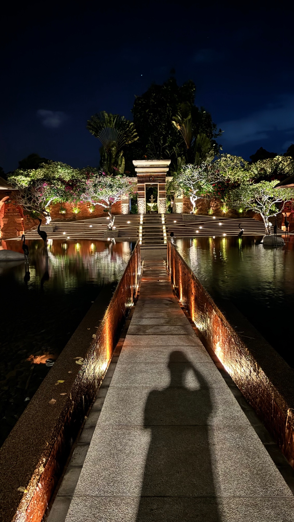 Stone causeway leading across a lit pond toward a Balinese gate at night