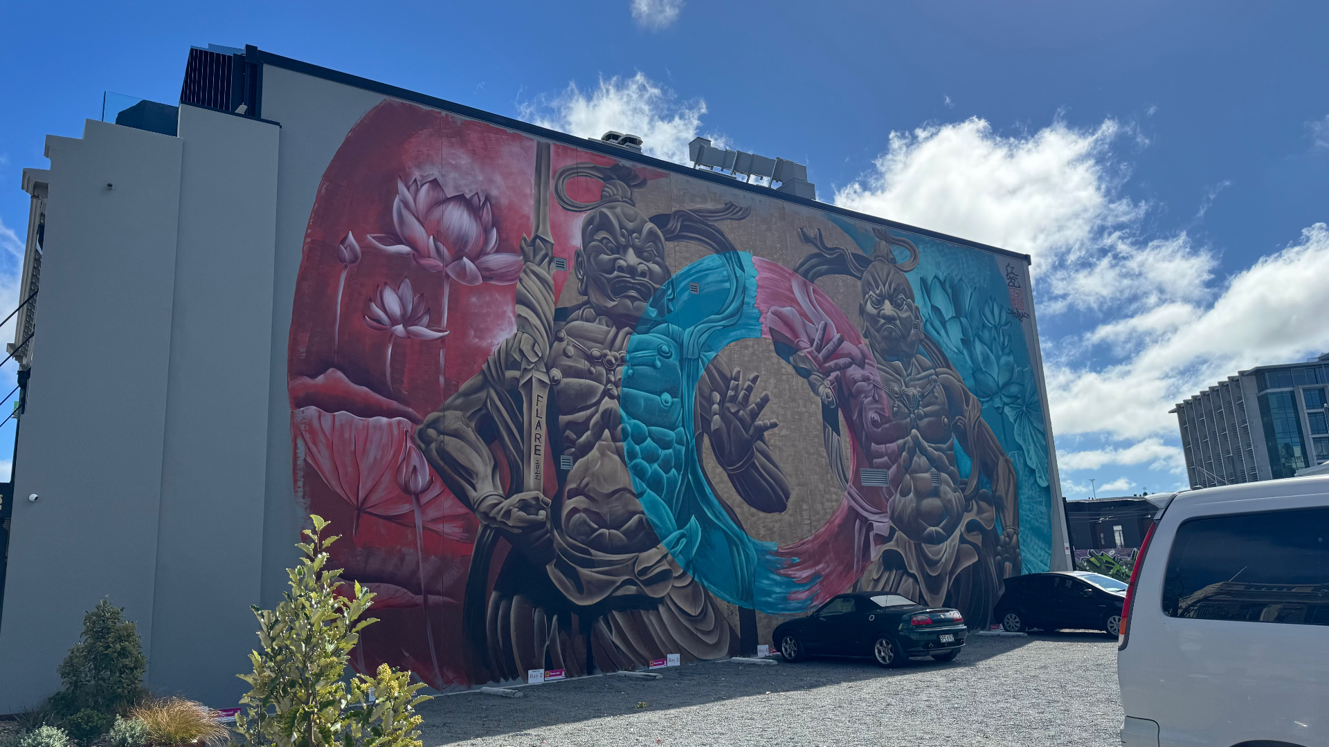 A large street mural of two guardian figures with lotus flowers in red and blue on the side of a downtown building