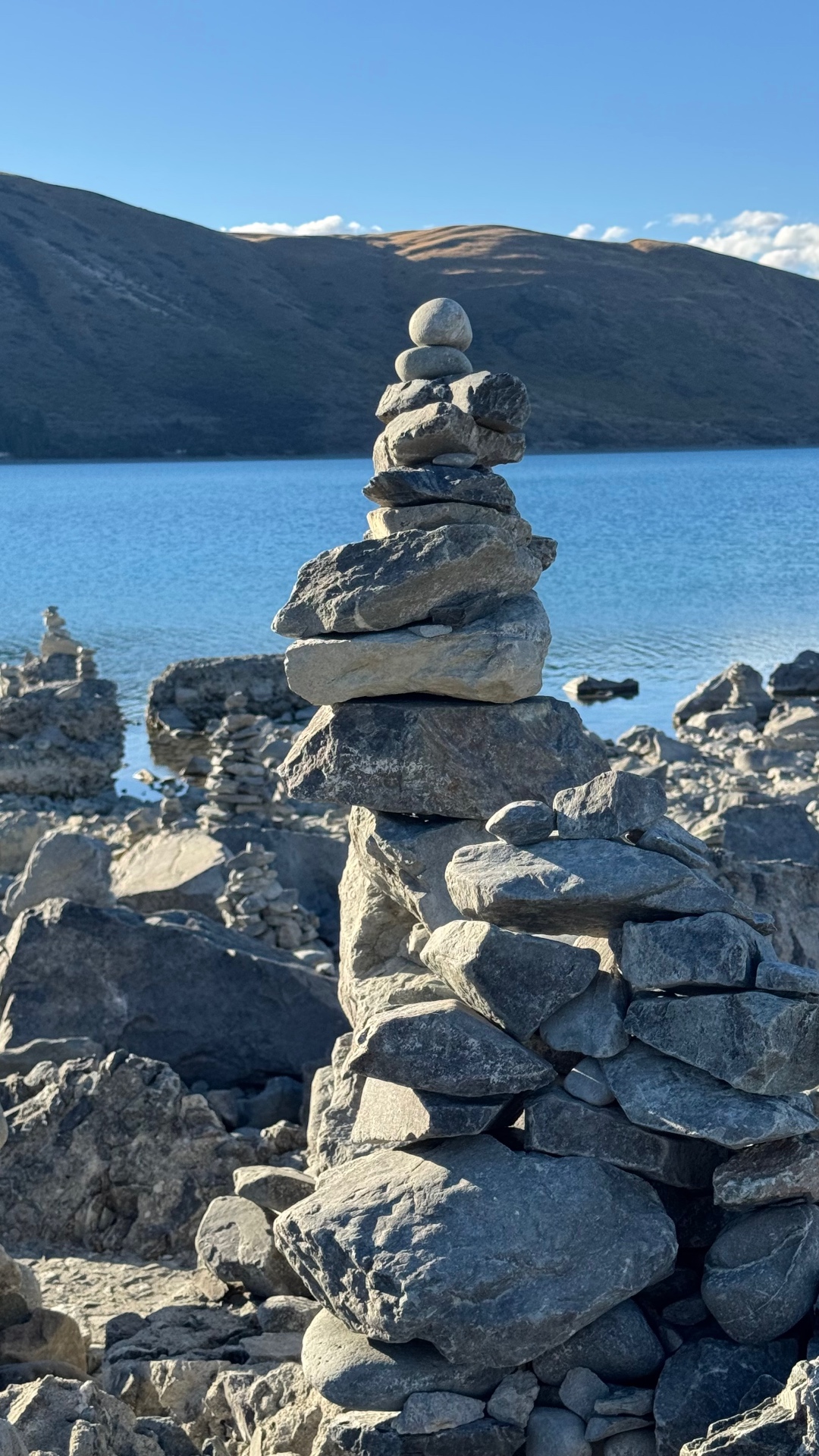 A tall stone cairn standing in front of the turquoise lake and a brown hillside
