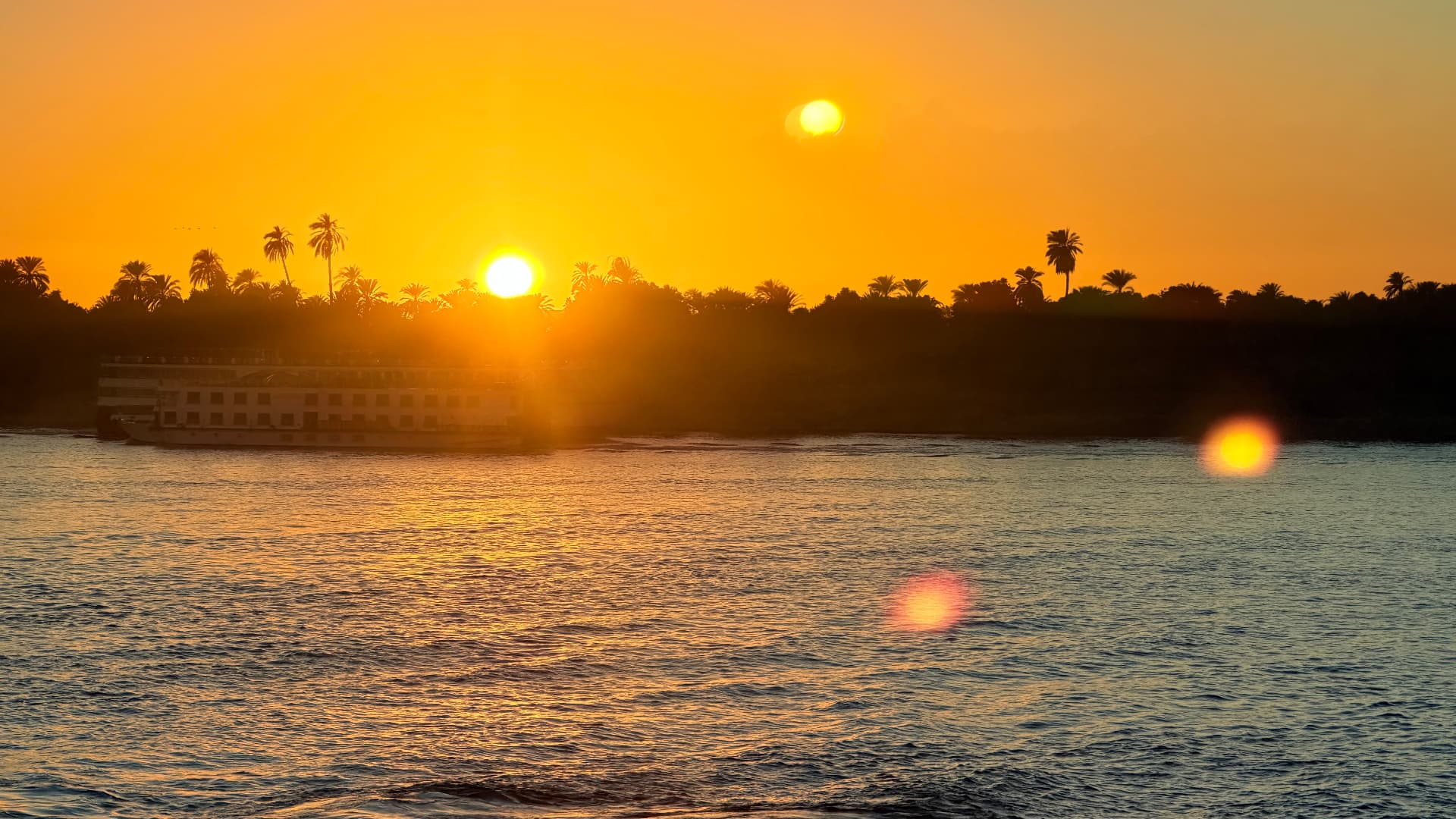 An orange Nile sunset behind palm silhouettes with a cruise boat passing along the far bank