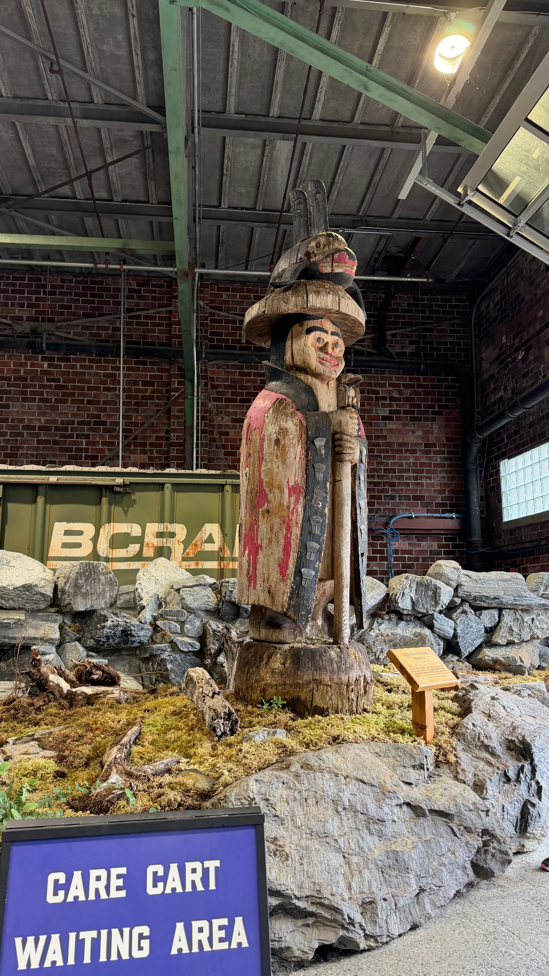 A carved and painted totem figure wearing a woven hat with a bird on top, holding a staff, displayed indoors on a mossy rock bed
