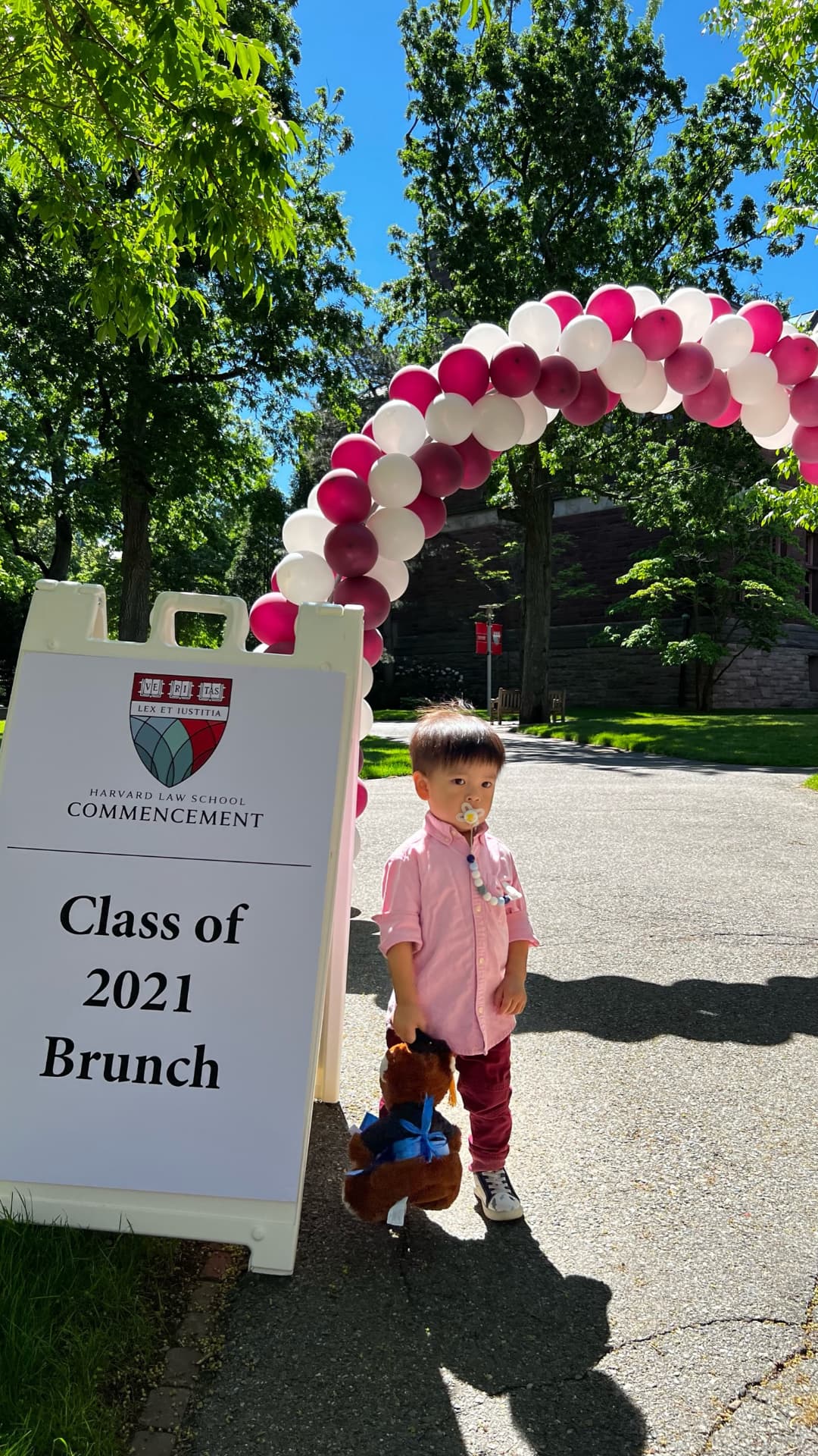 A little boy in pink beside the Harvard Law School Commencement Class of 2021 Brunch sign and balloon arch