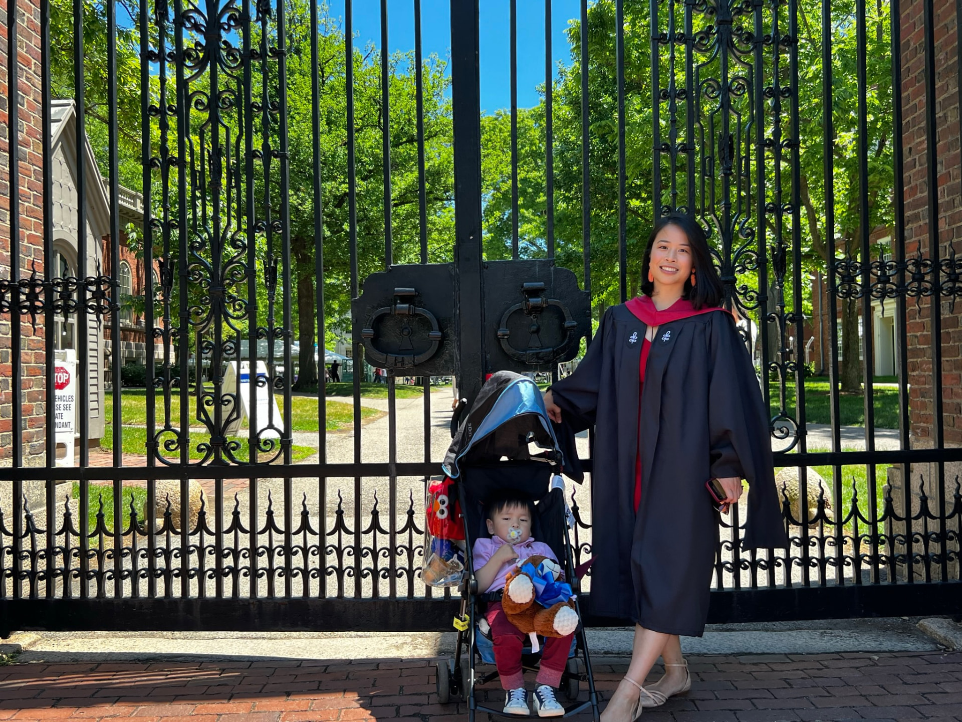 Jocelyn in her graduation gown at the wrought-iron gates of Harvard Yard, with a little one in a stroller