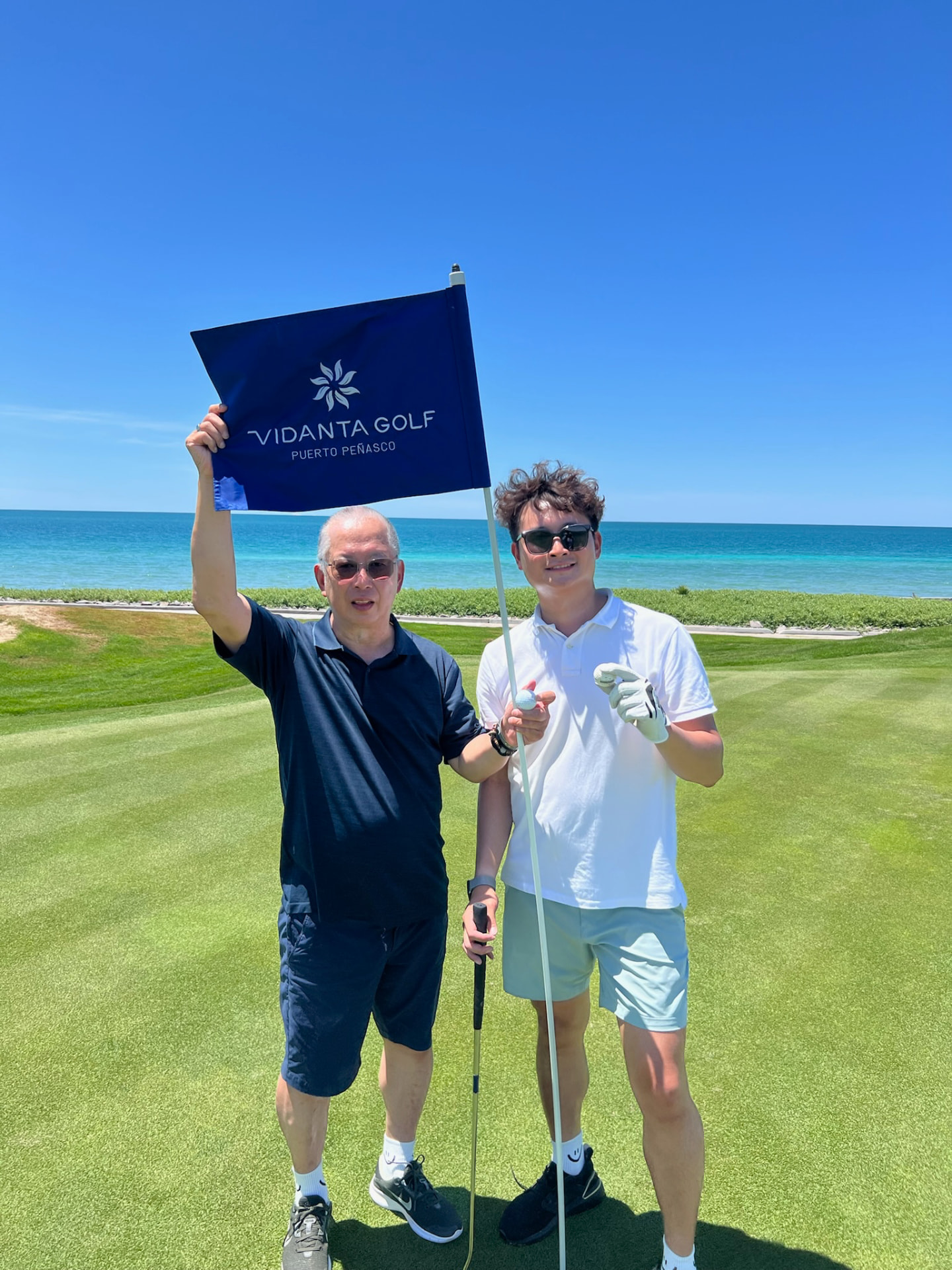Ken and Johan holding a Vidanta Golf Puerto Peñasco flag beside the sea
